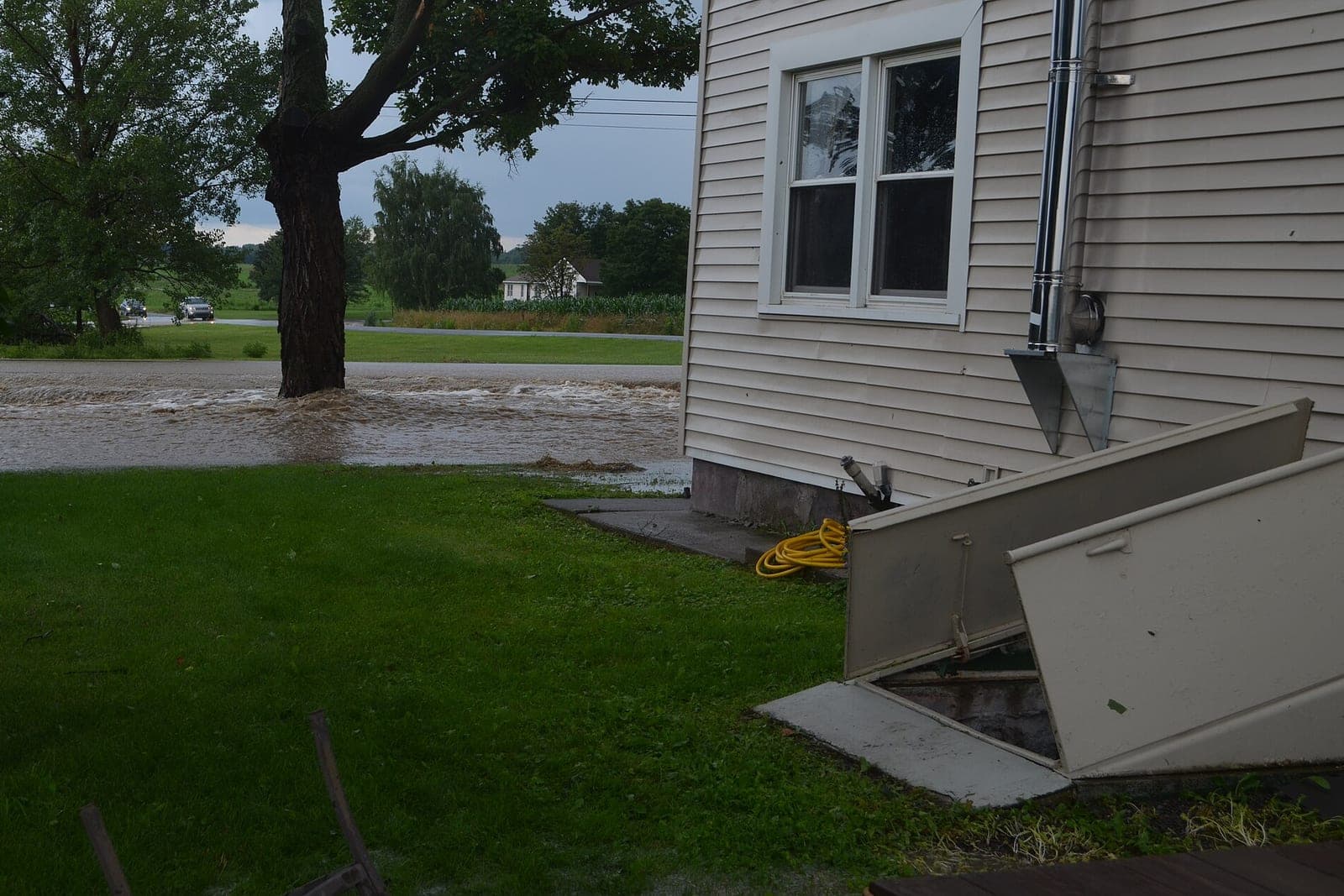 Brown floodwater runs across a lawn and road beside a house with an open cellar door