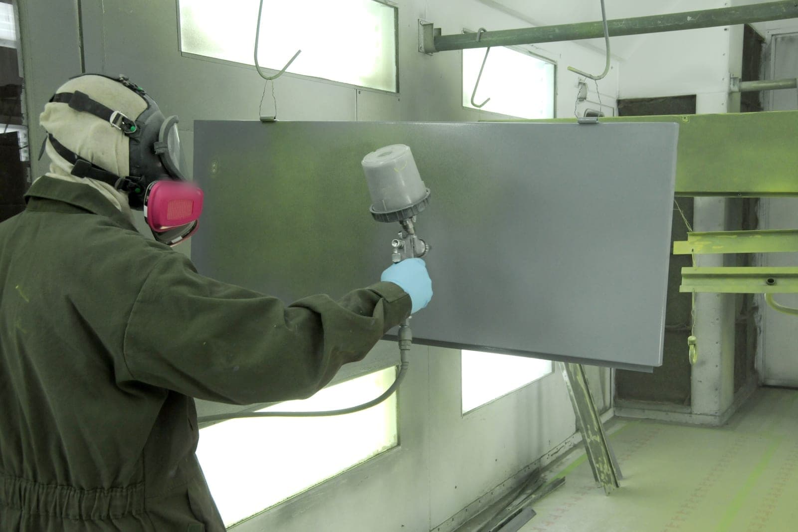 A painter in a hood and respirator sprays a gray coat on a large panel hanging inside a paint booth