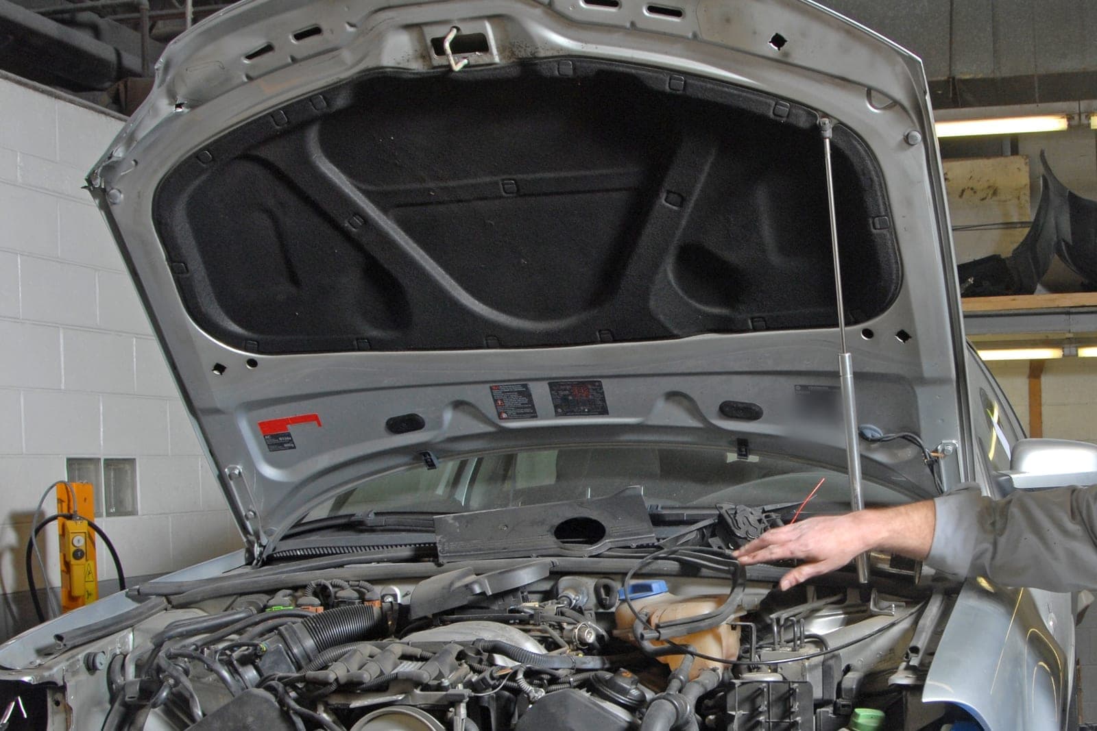 A silver car with its hood raised and front bumper cover off sits in a repair bay while bumper covers wait on a shelf