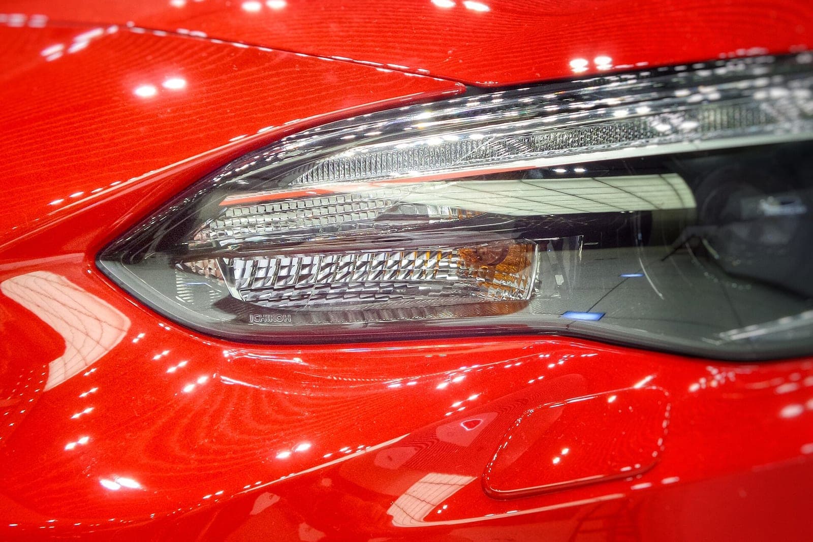 A close view of a glossy red car fender and headlight reflecting rows of shop lights