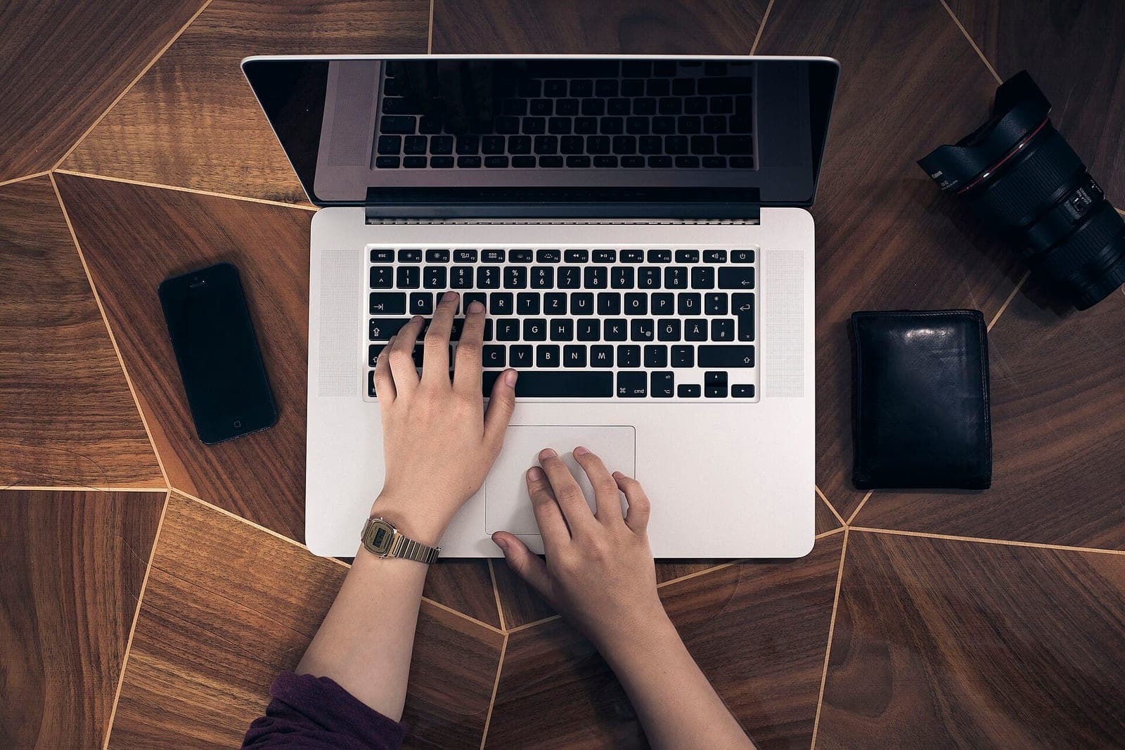 Hands type on a laptop on a wooden table, with a phone, a wallet and a camera lens nearby
