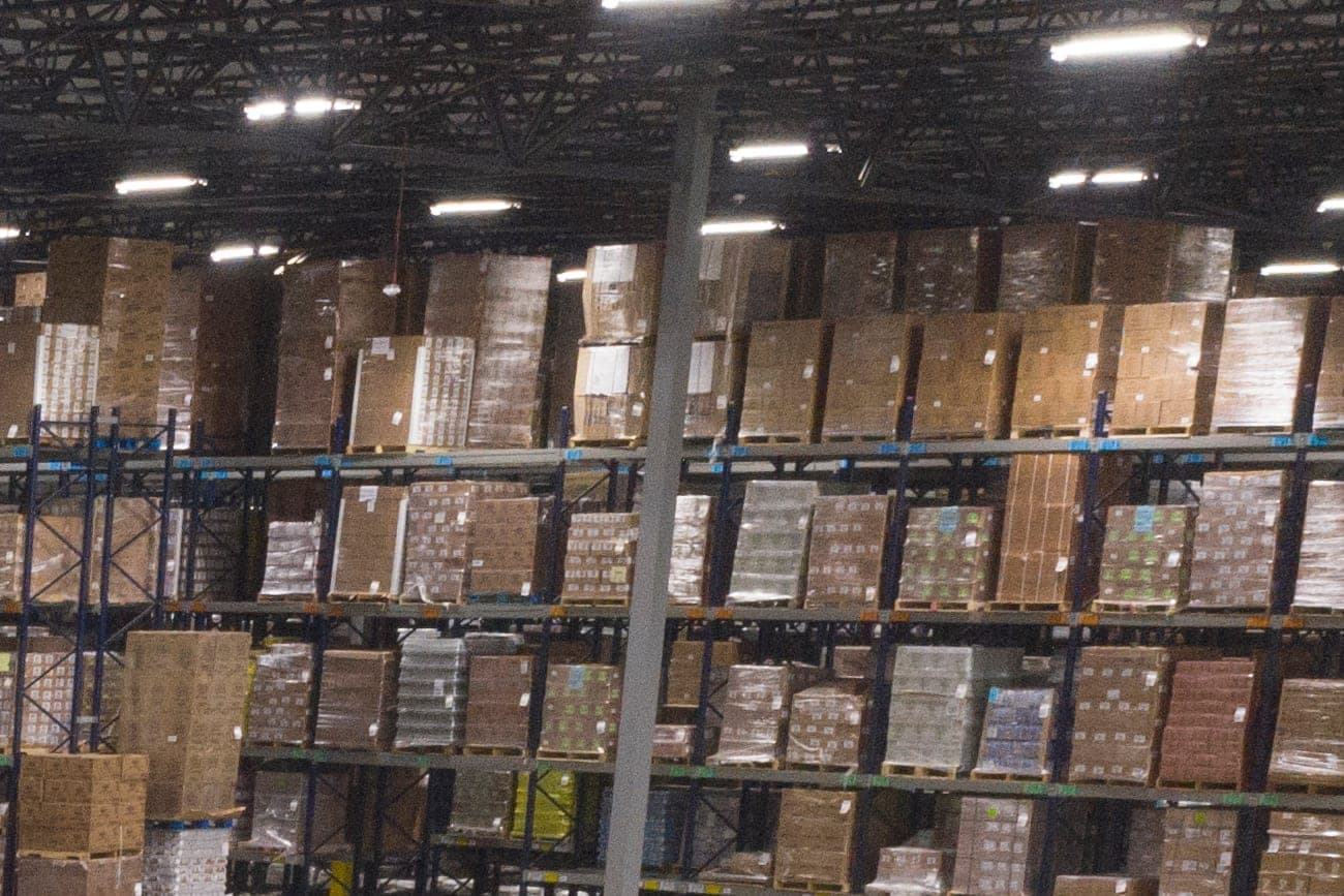Rows of steel warehouse racks stacked high with wrapped pallets of cartons under bright ceiling lights