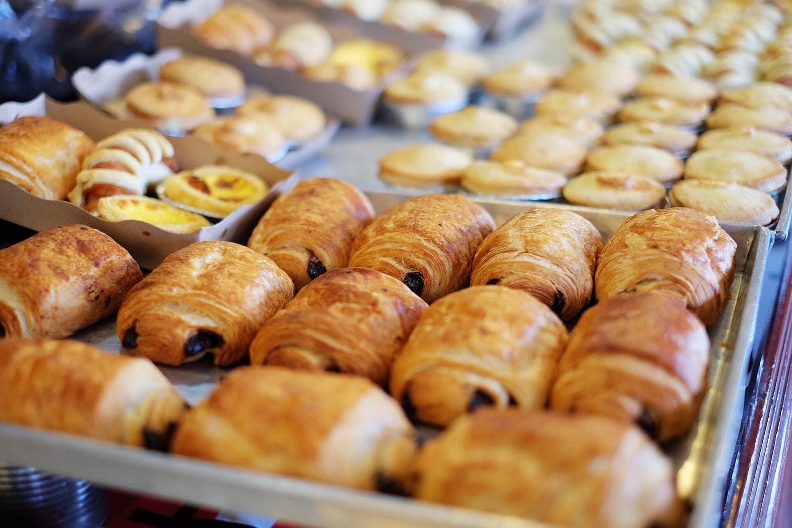 Metal trays of chocolate-filled croissants and small custard tarts lined up on a bakery counter