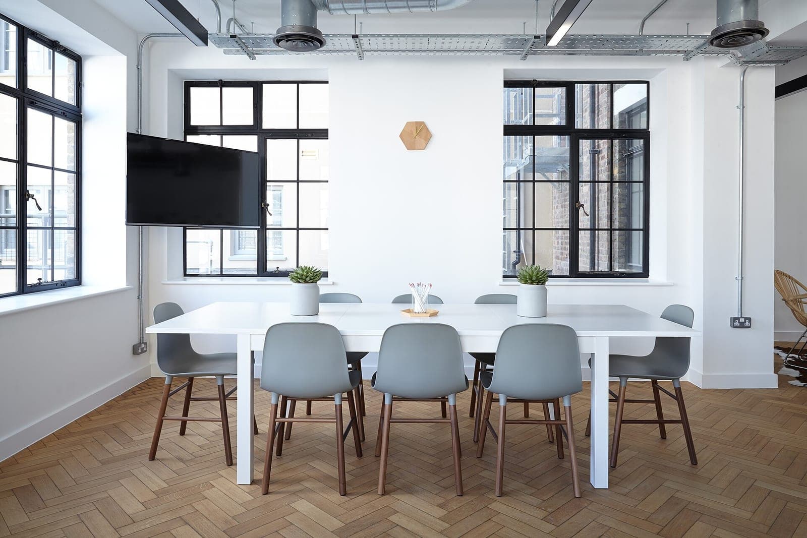 An empty meeting room with a long white table, gray chairs, two potted plants and tall black-framed windows