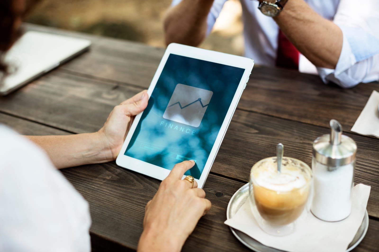 A person holds a tablet showing a finance chart icon at a wooden table beside a cup of coffee