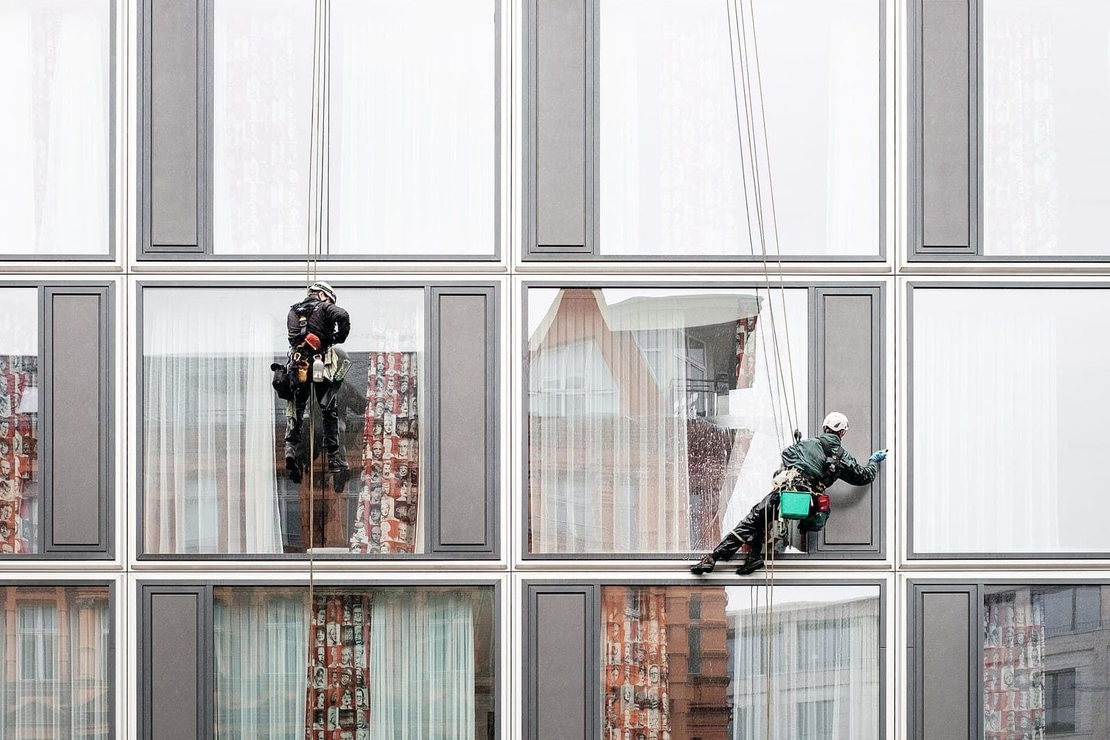 Two window cleaners in helmets hang from ropes on the glass front of a modern building