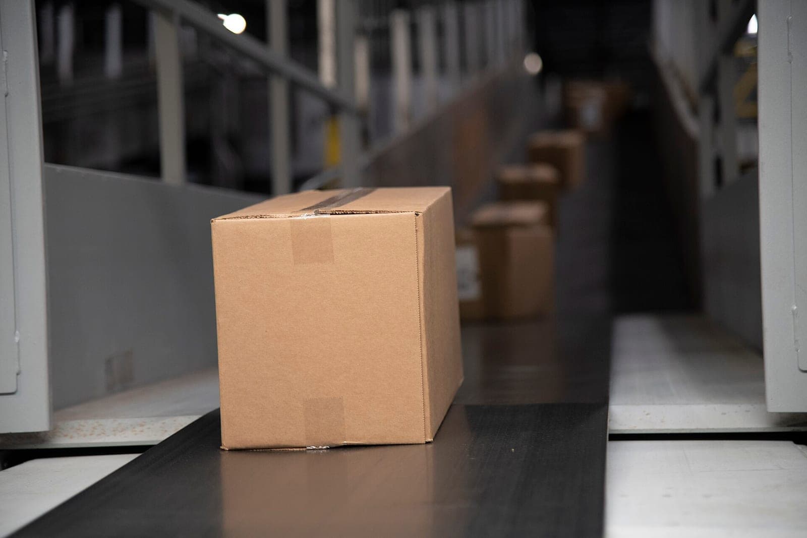A plain cardboard box moves along a dark conveyor belt, with more boxes blurred behind it