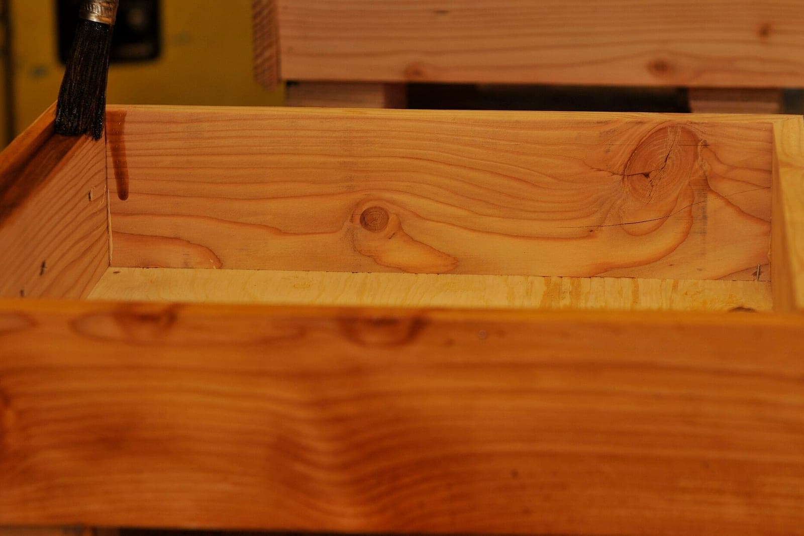A dark brush rests on the edge of a pine box while wood stain runs down its inside wall