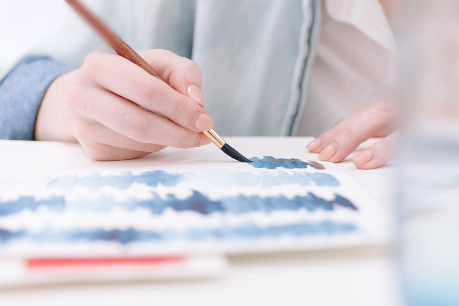 A hand paints rows of blue watercolor strokes on white paper with a fine brush