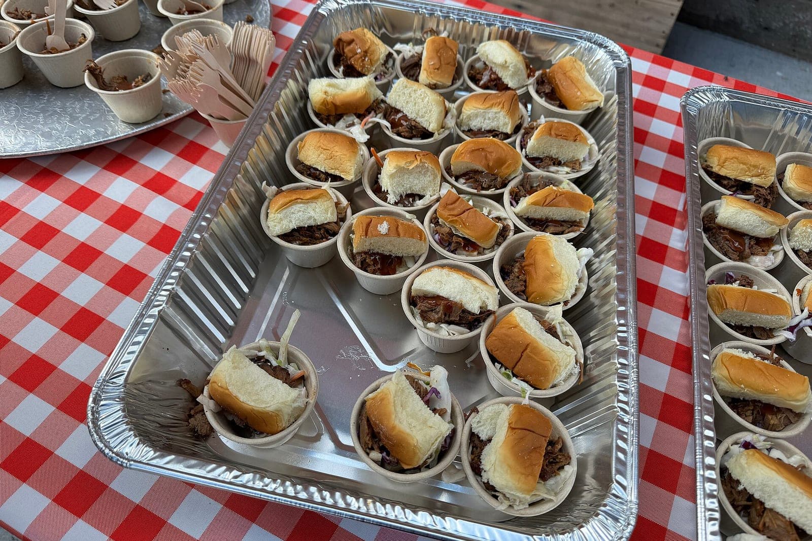 Foil catering pans of pulled meat sliders with slaw in paper cups on a red and white checked tablecloth