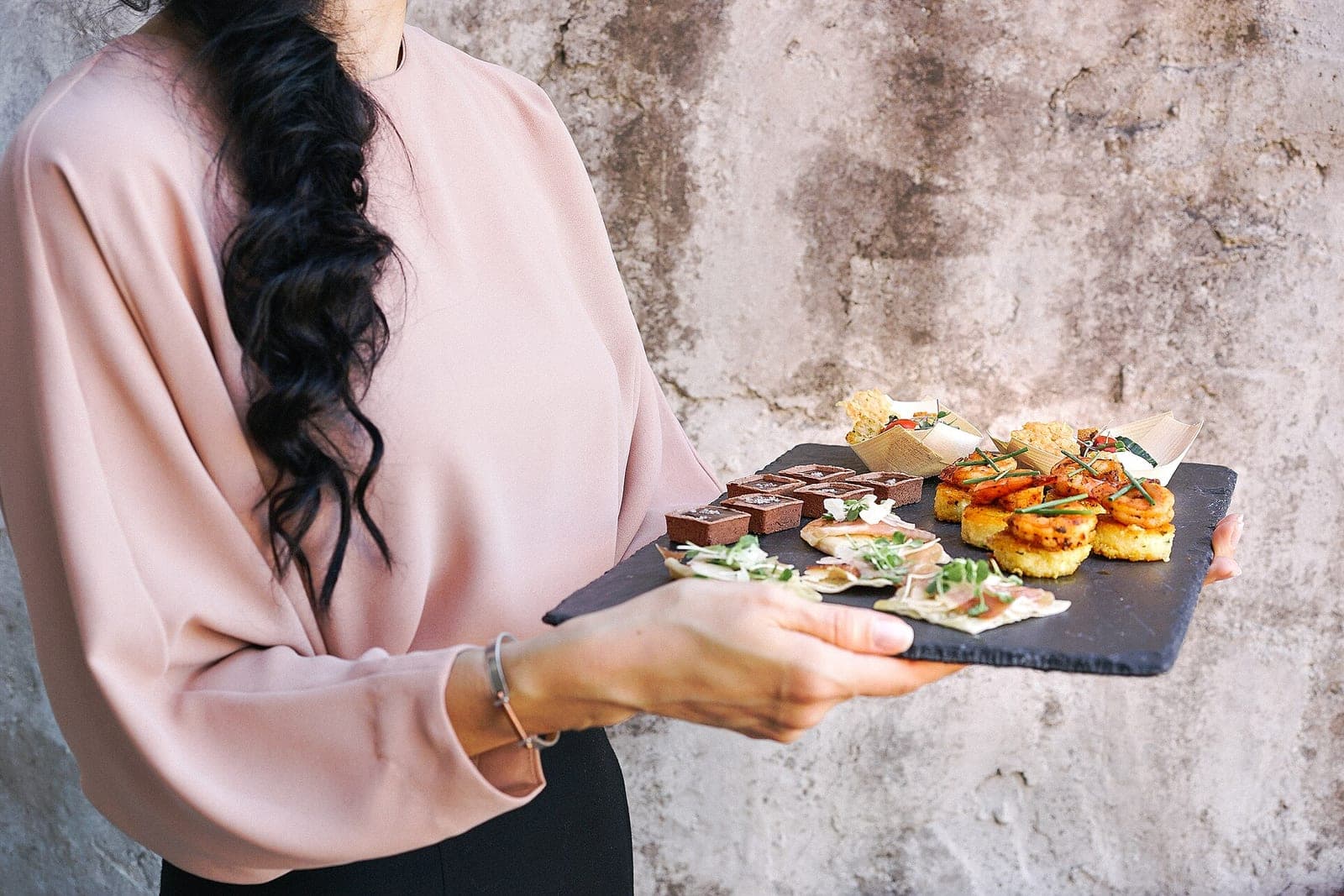 A server in a pink blouse, seen from the shoulders down, holding a slate tray of shrimp bites, small tarts and crackers