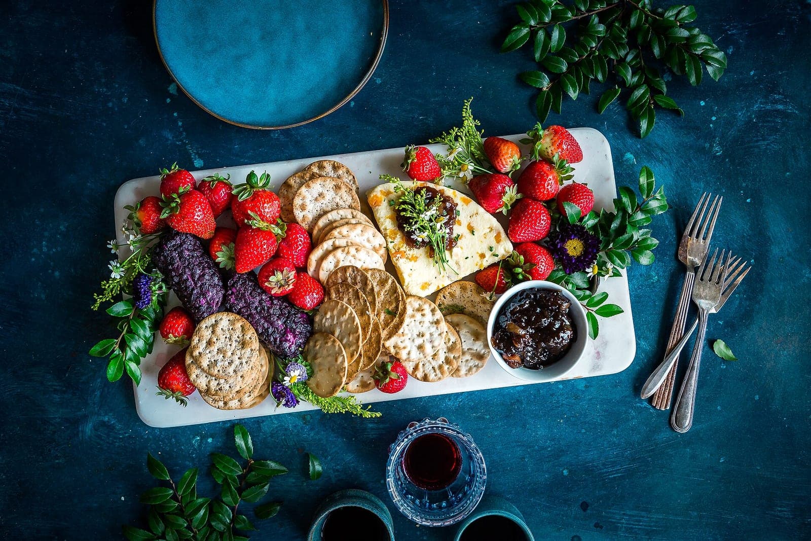 A white serving board of crackers, strawberries, cheese and jam sits on a dark blue table beside two forks