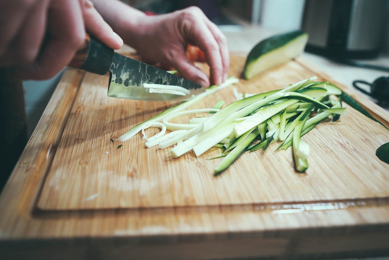 Two hands slicing a zucchini into thin strips with a chef's knife on a wooden cutting board