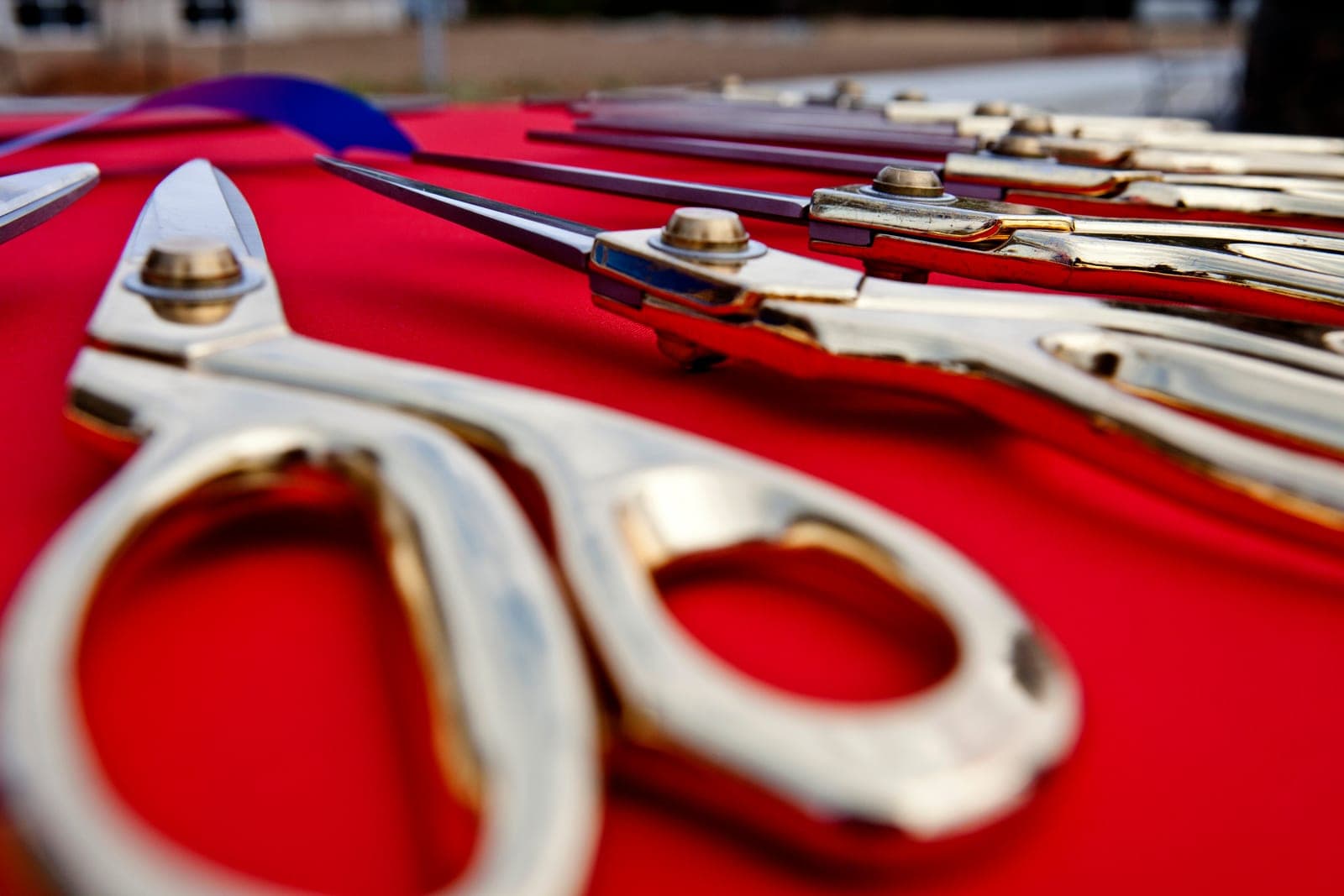 Rows of large gold ceremonial scissors rest on a red tablecloth beside a blue ribbon