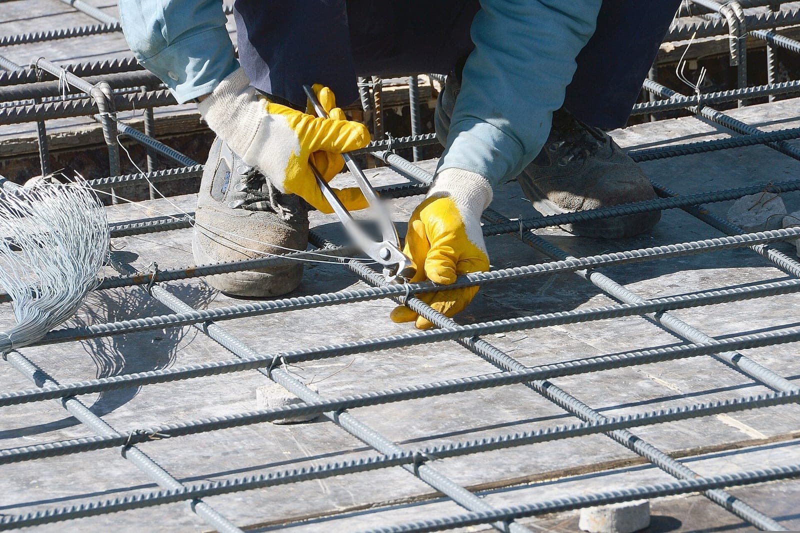 Gloved hands tie steel reinforcing bars together with pliers over wooden formwork for a concrete floor