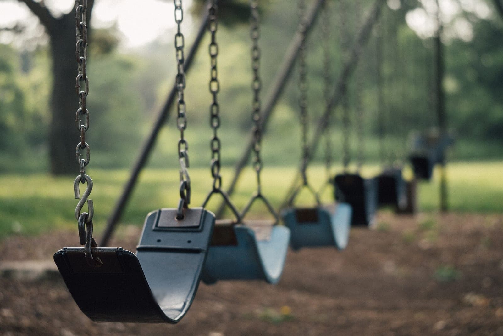 A row of empty blue swing seats hanging on chains over a dirt play area, with green trees behind