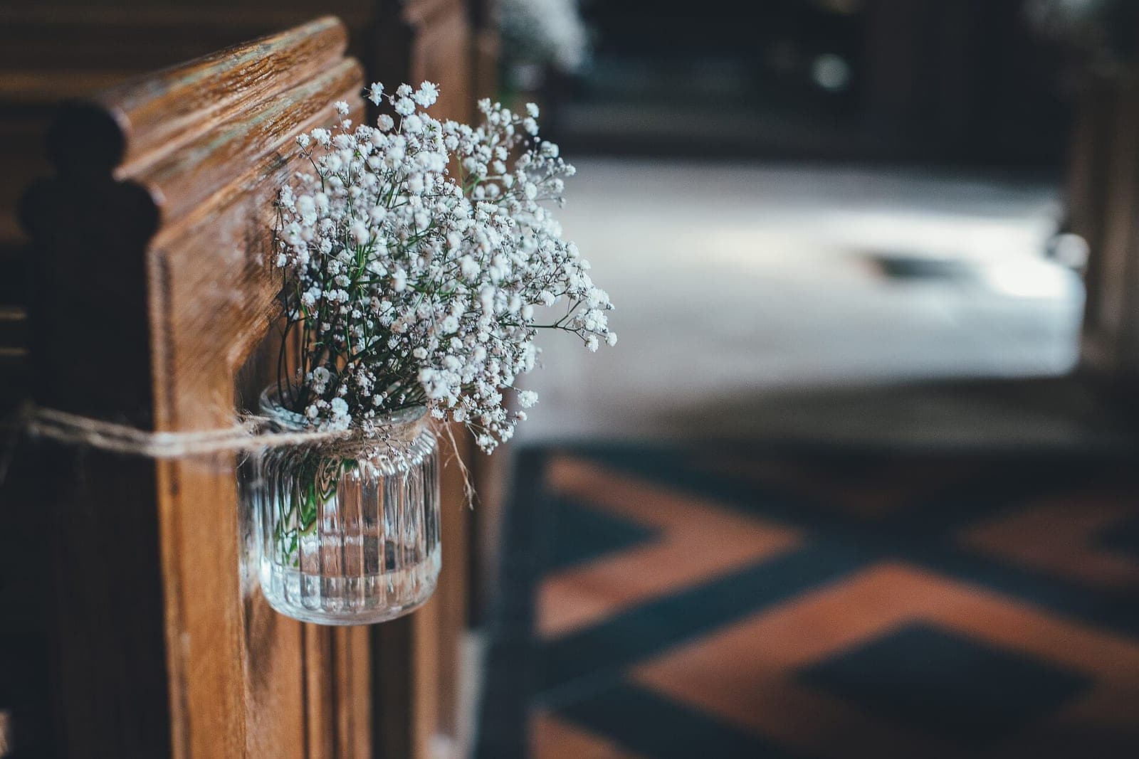 A small jar of white baby's breath tied with twine to the end of a wooden church pew beside a tiled aisle