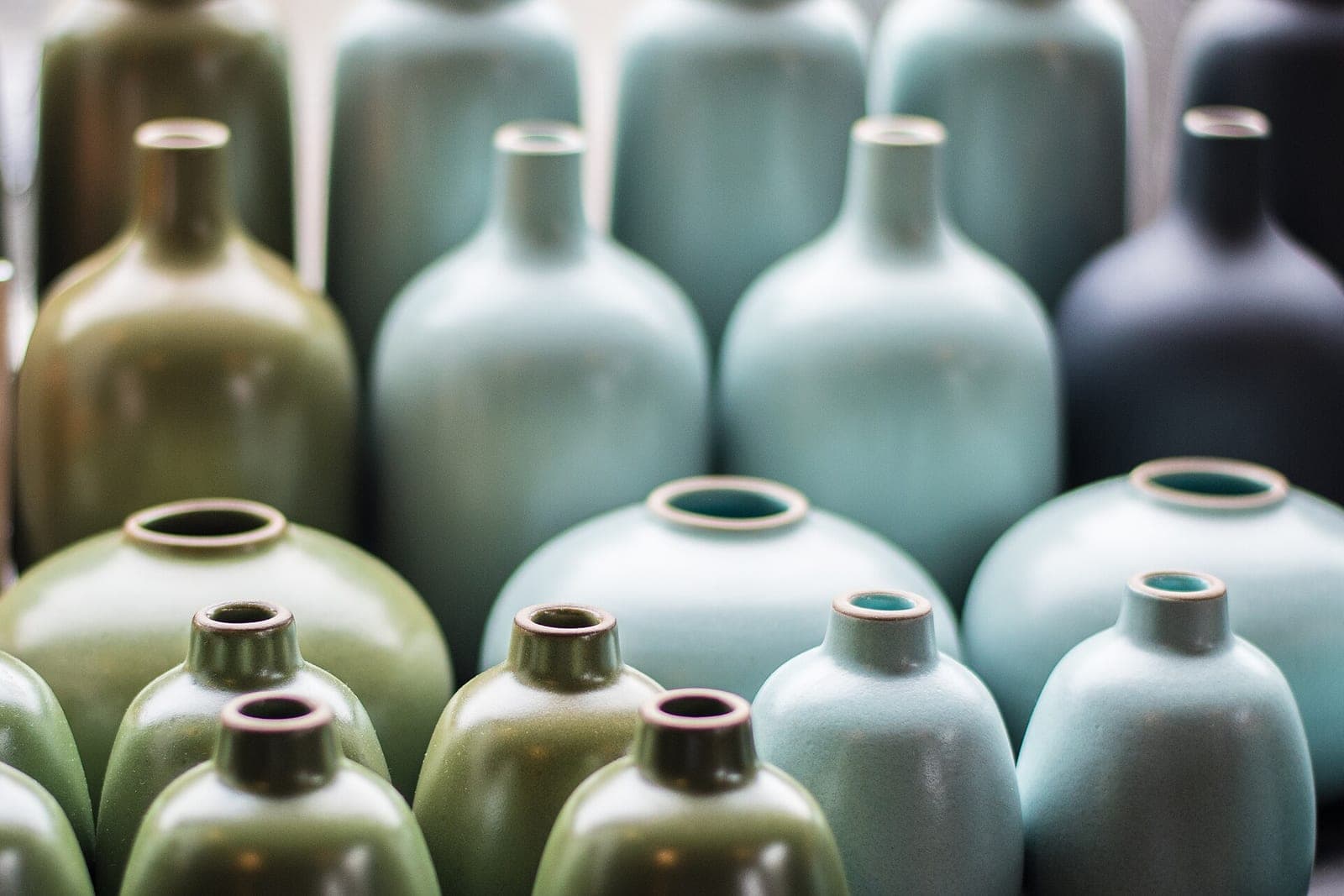Rows of glazed ceramic bottles and round vases in green, pale blue and dark gray glazes