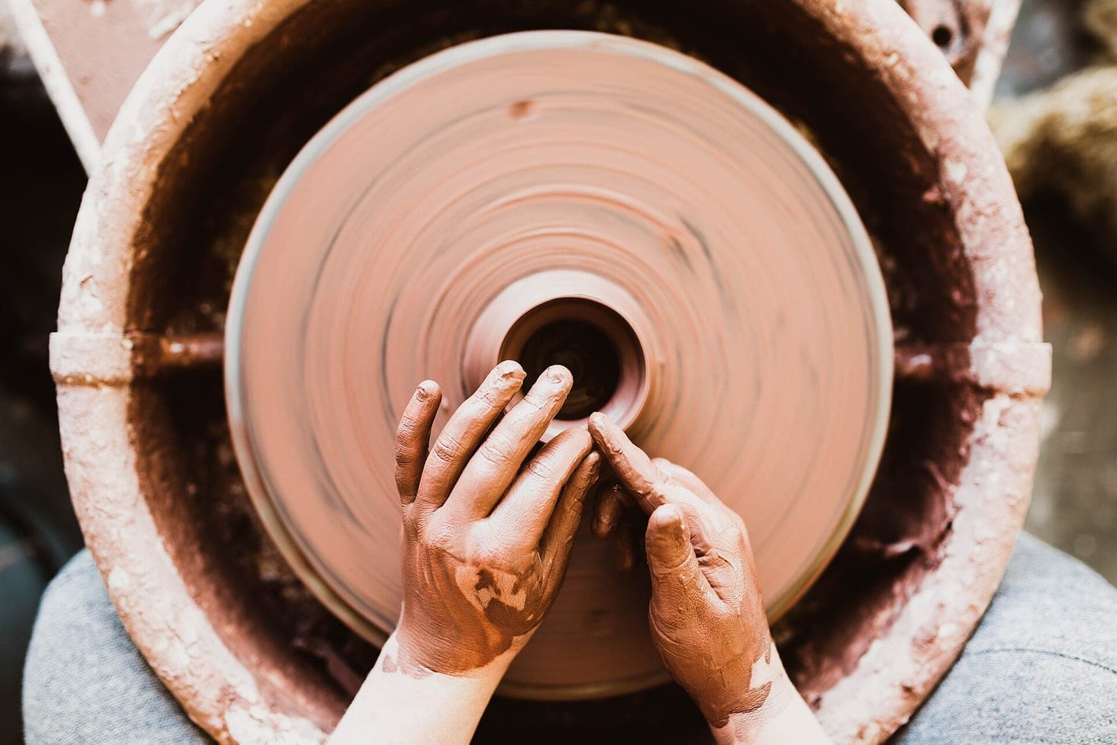 Clay-covered hands shape a small pot at the center of a spinning pottery wheel