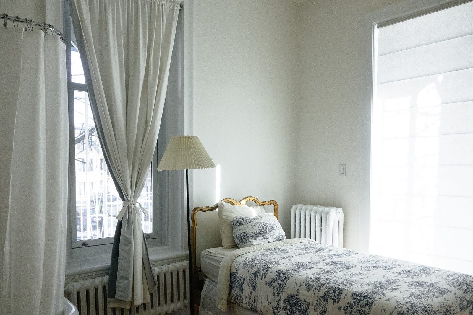A neatly made single bed with a patterned cover beside a floor lamp and tall curtained window