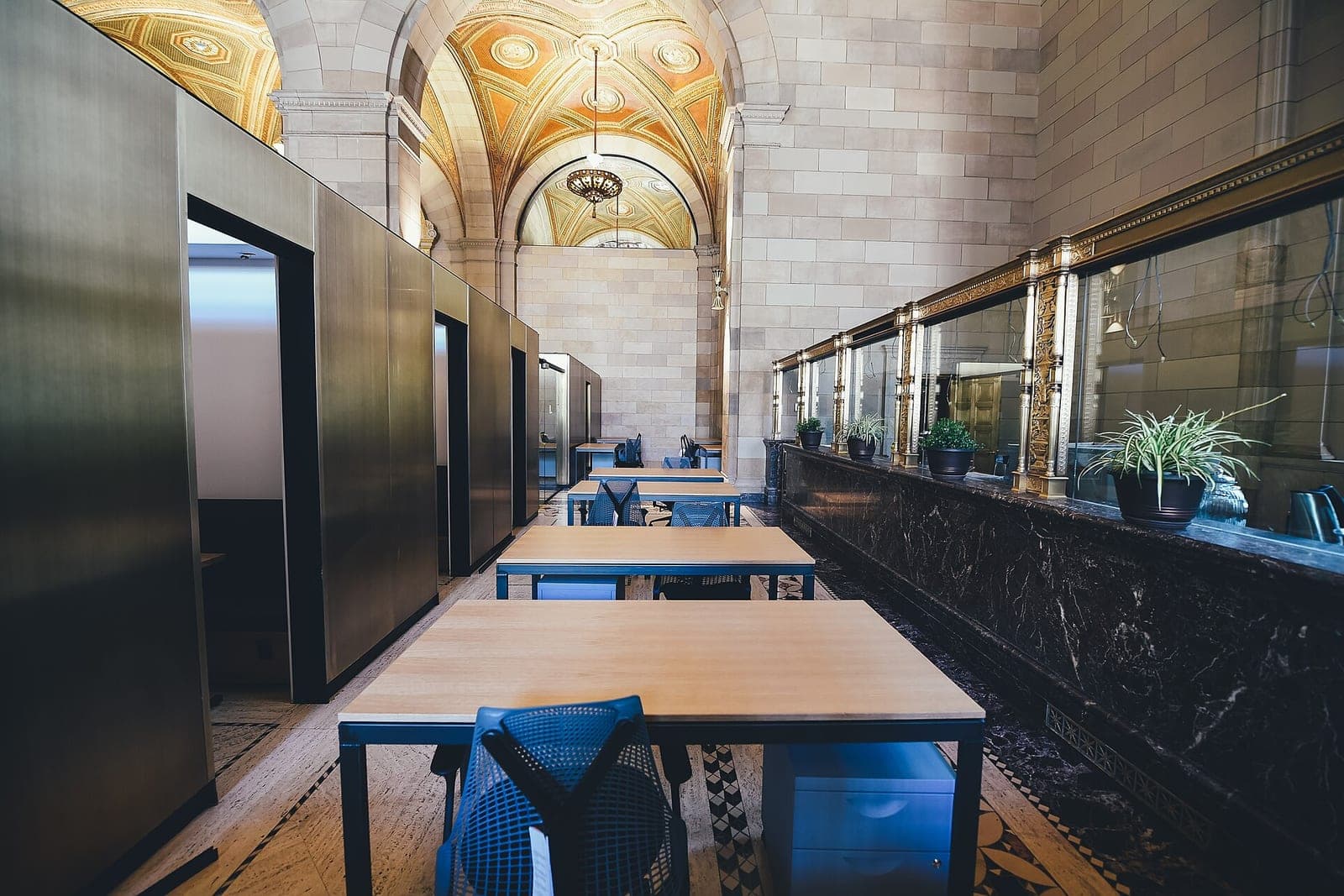 Wooden desks with blue chairs in a long office hall beside a marble counter with potted plants