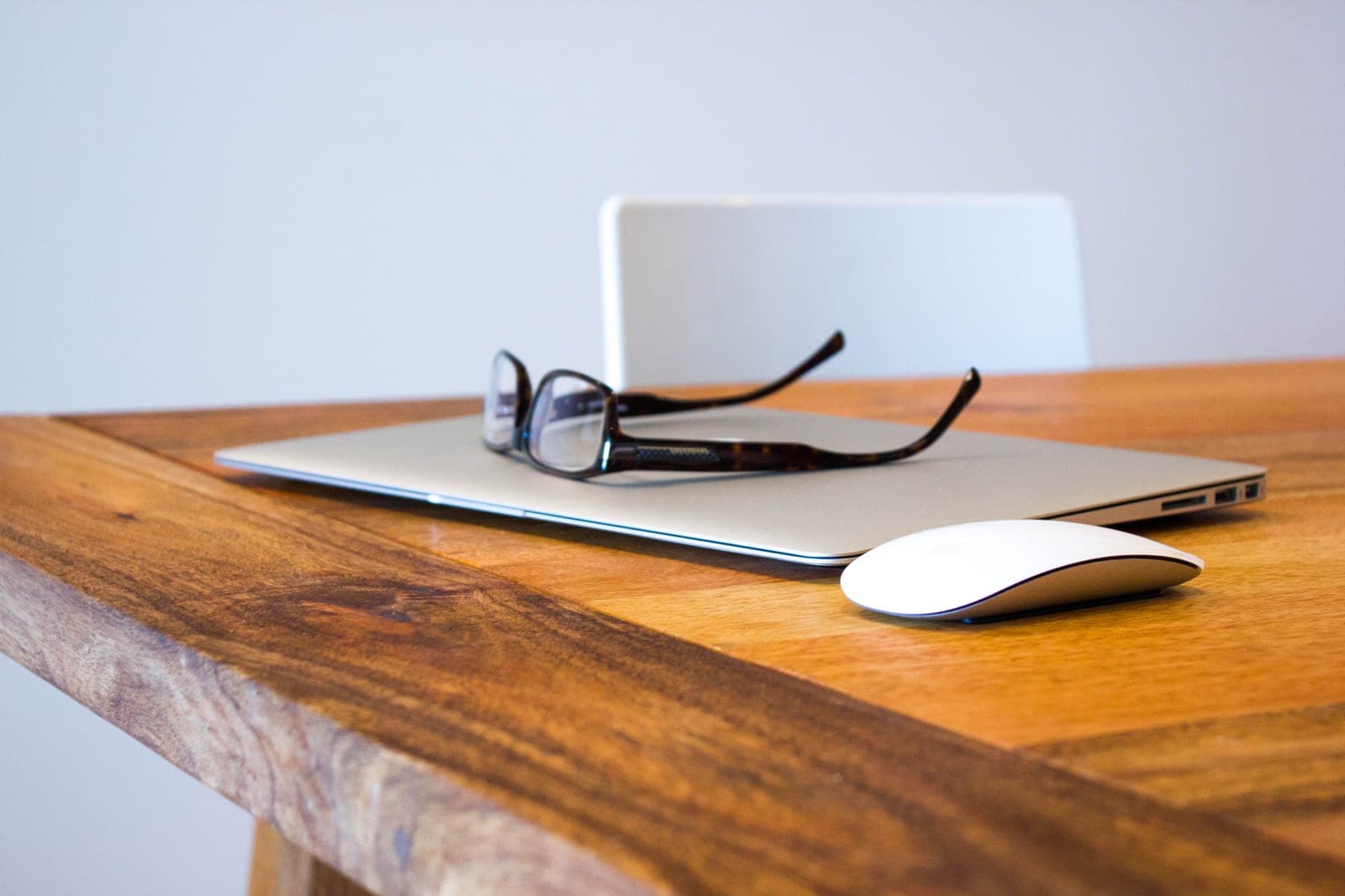 Reading glasses rest on a closed laptop beside a white mouse on a wooden desk