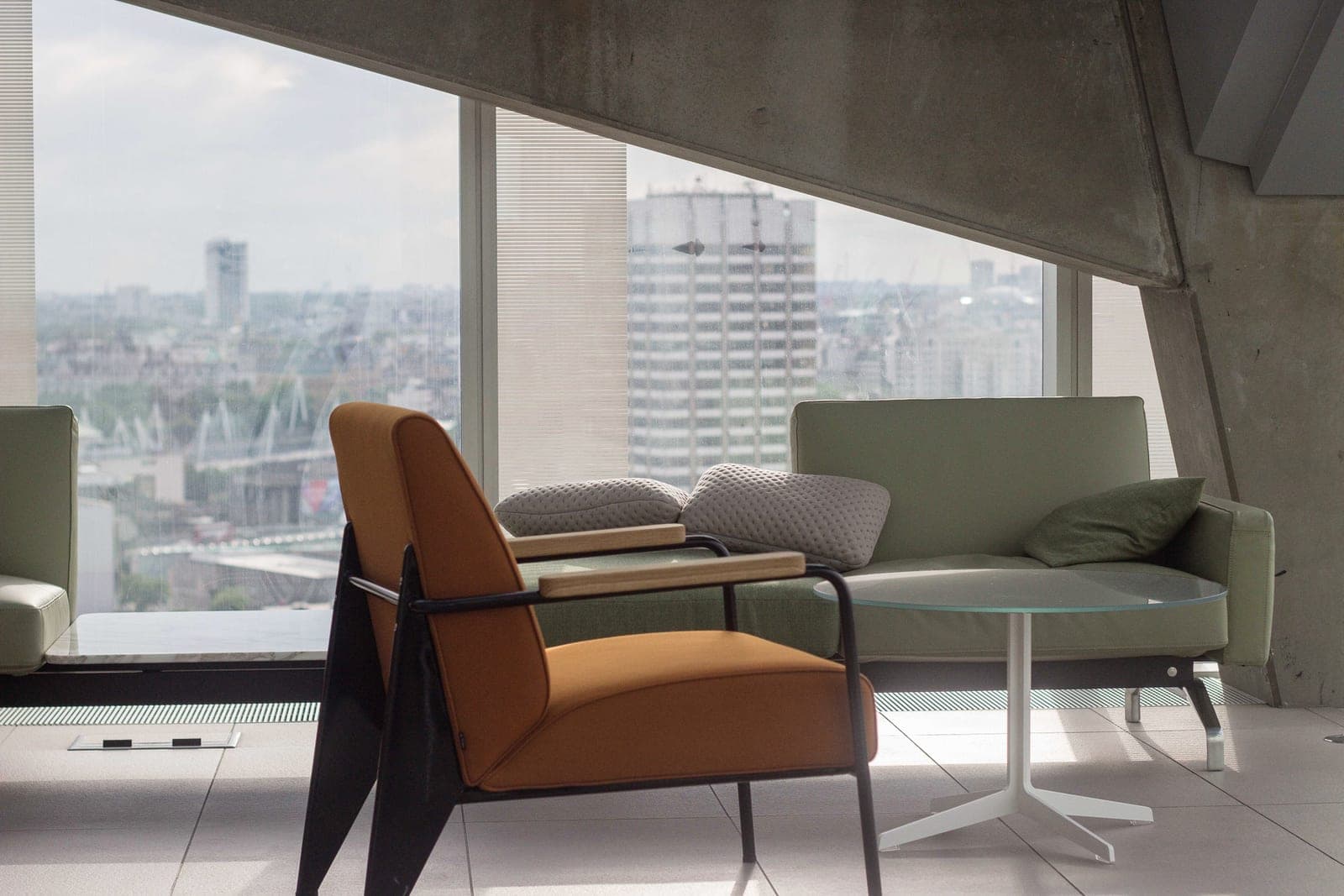 An orange armchair, a green sofa and a round glass table sit by tall windows over a city