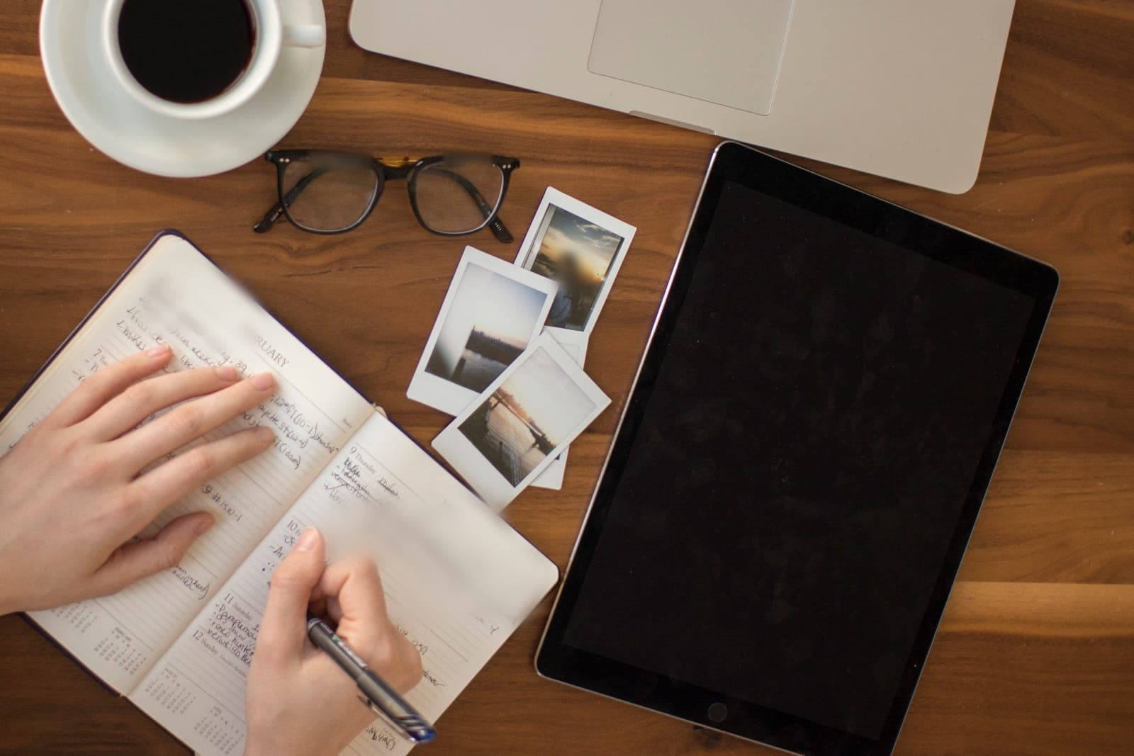 Hands write in an open planner beside a coffee cup, reading glasses, instant photos and a tablet
