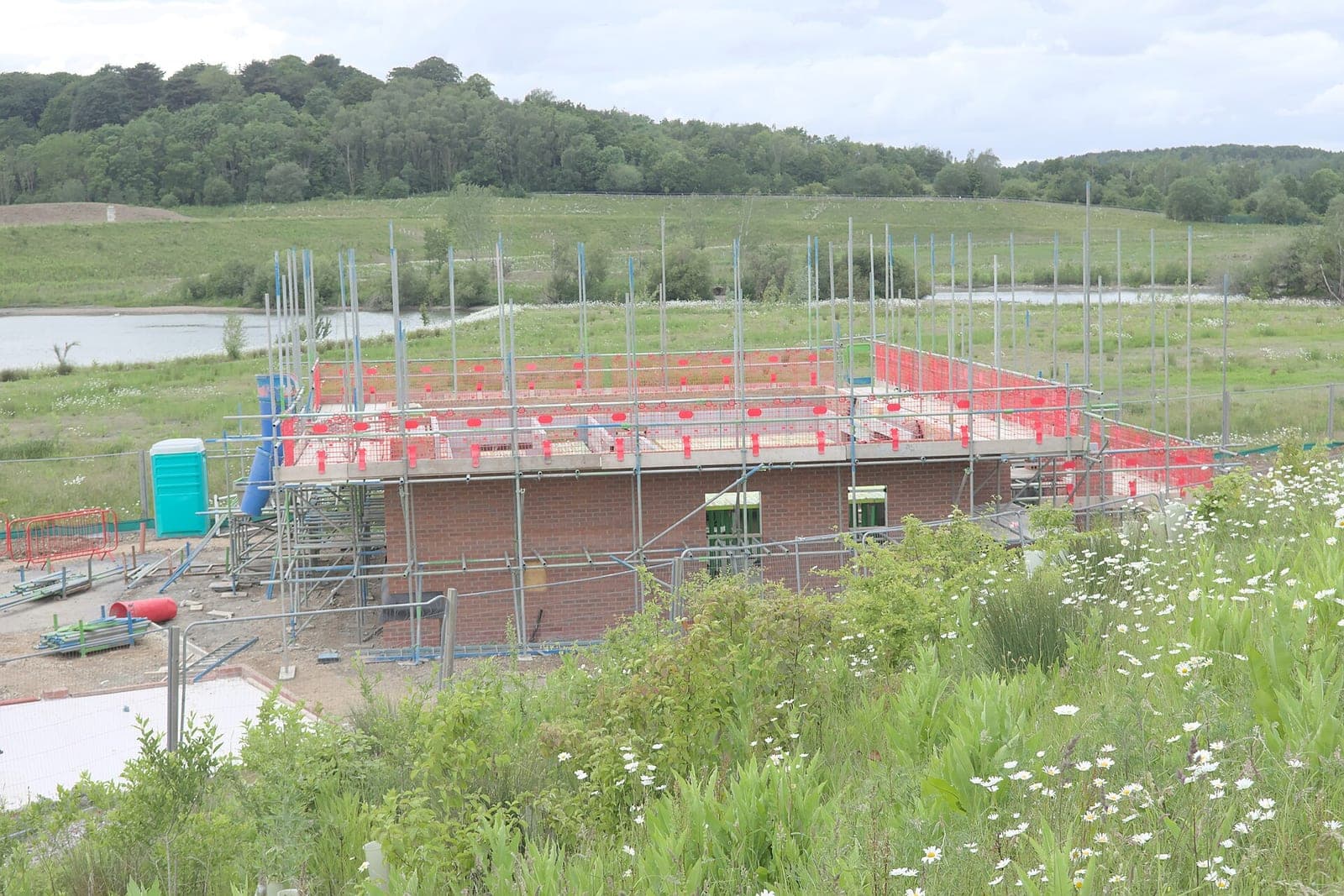 The brick ground floor of a new house wrapped in scaffolding, with a pond and green hills behind it