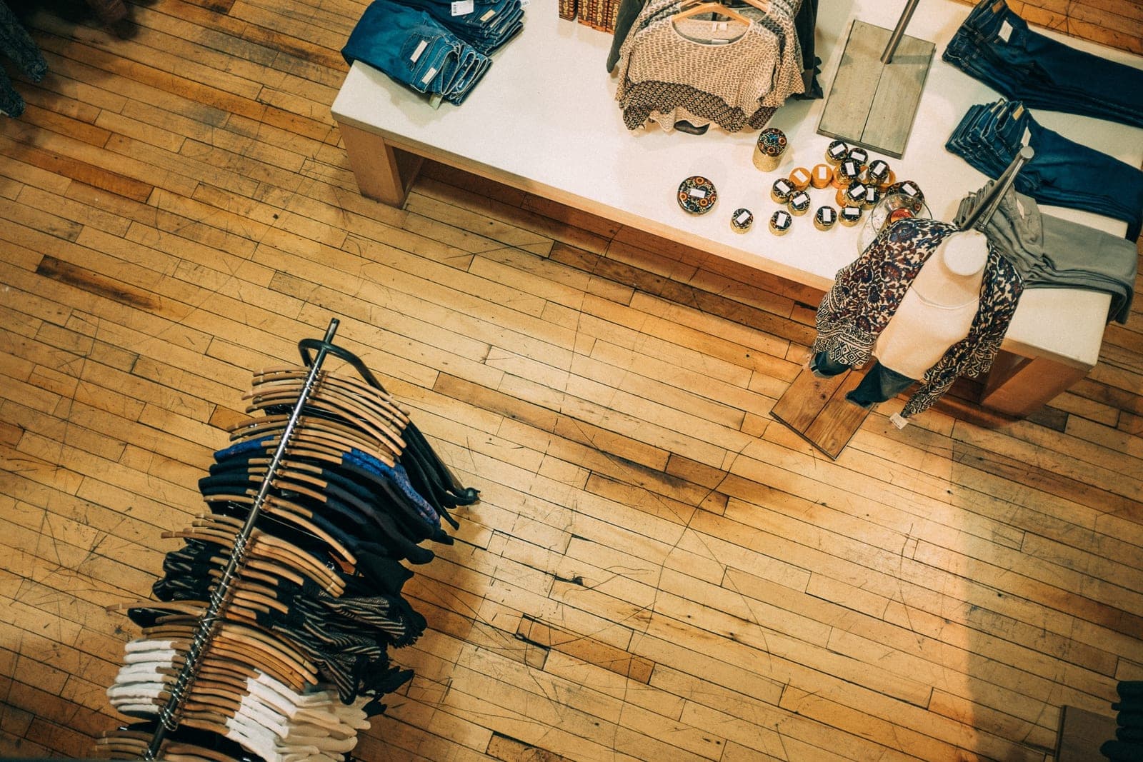 Seen from above, a rack of shirts on wooden hangers stands near a white table of folded jeans and small goods on a worn wood floor