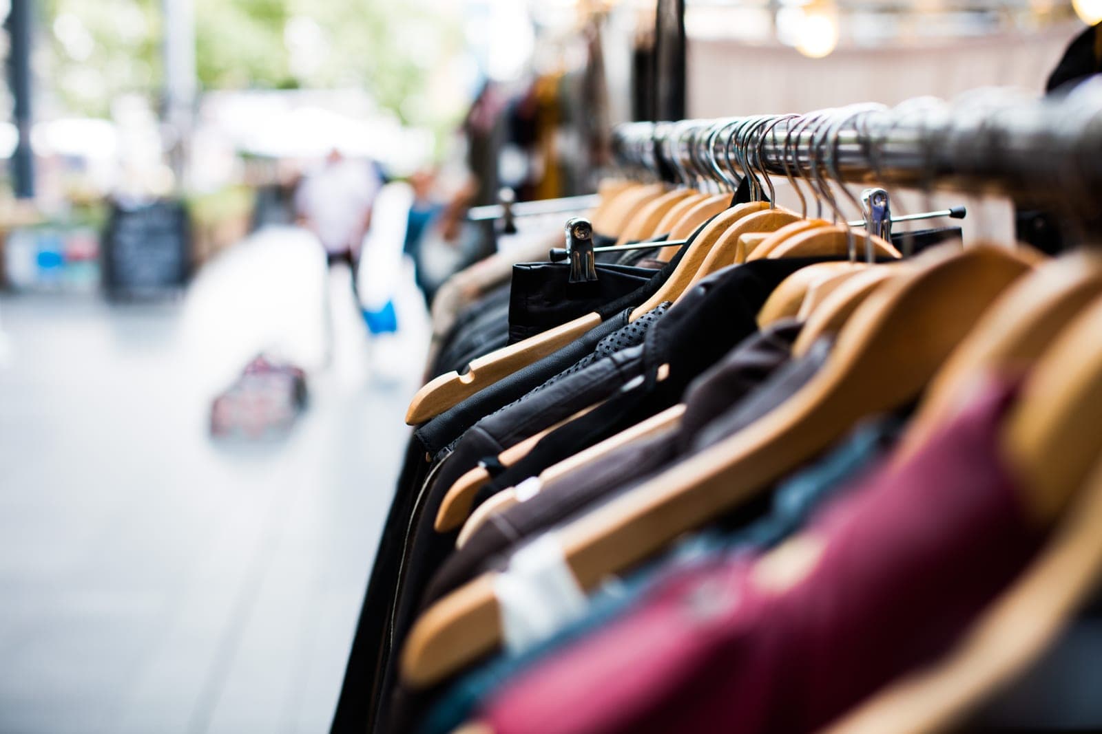 Jackets and sweaters hang on wooden hangers along a metal rack beside a blurred shopping street