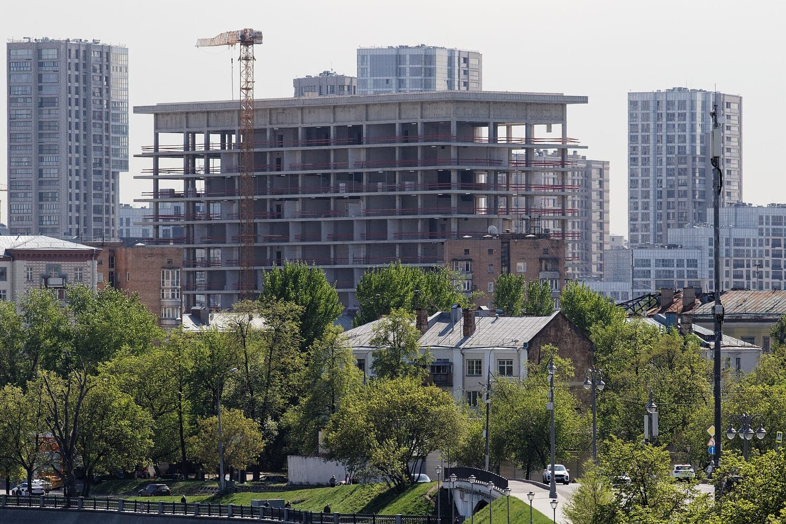 A concrete frame building under construction with a tower crane, seen above trees and older homes