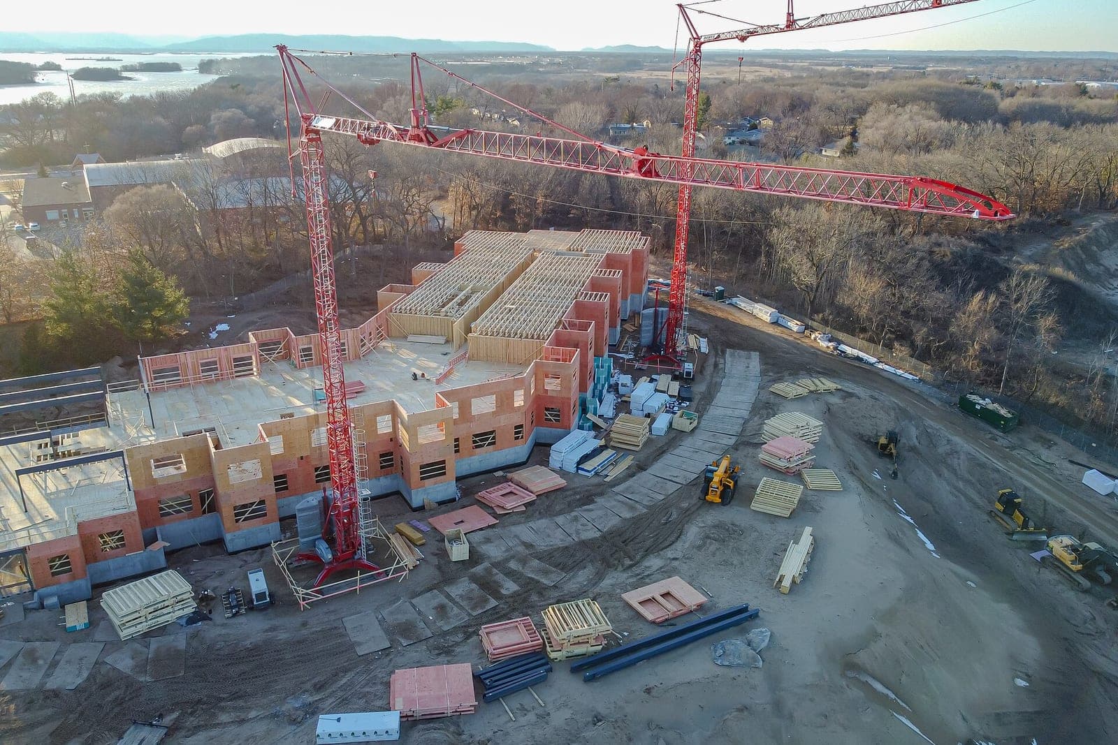 An aerial view of a large wood-framed building going up beside two red tower cranes and stacks of materials