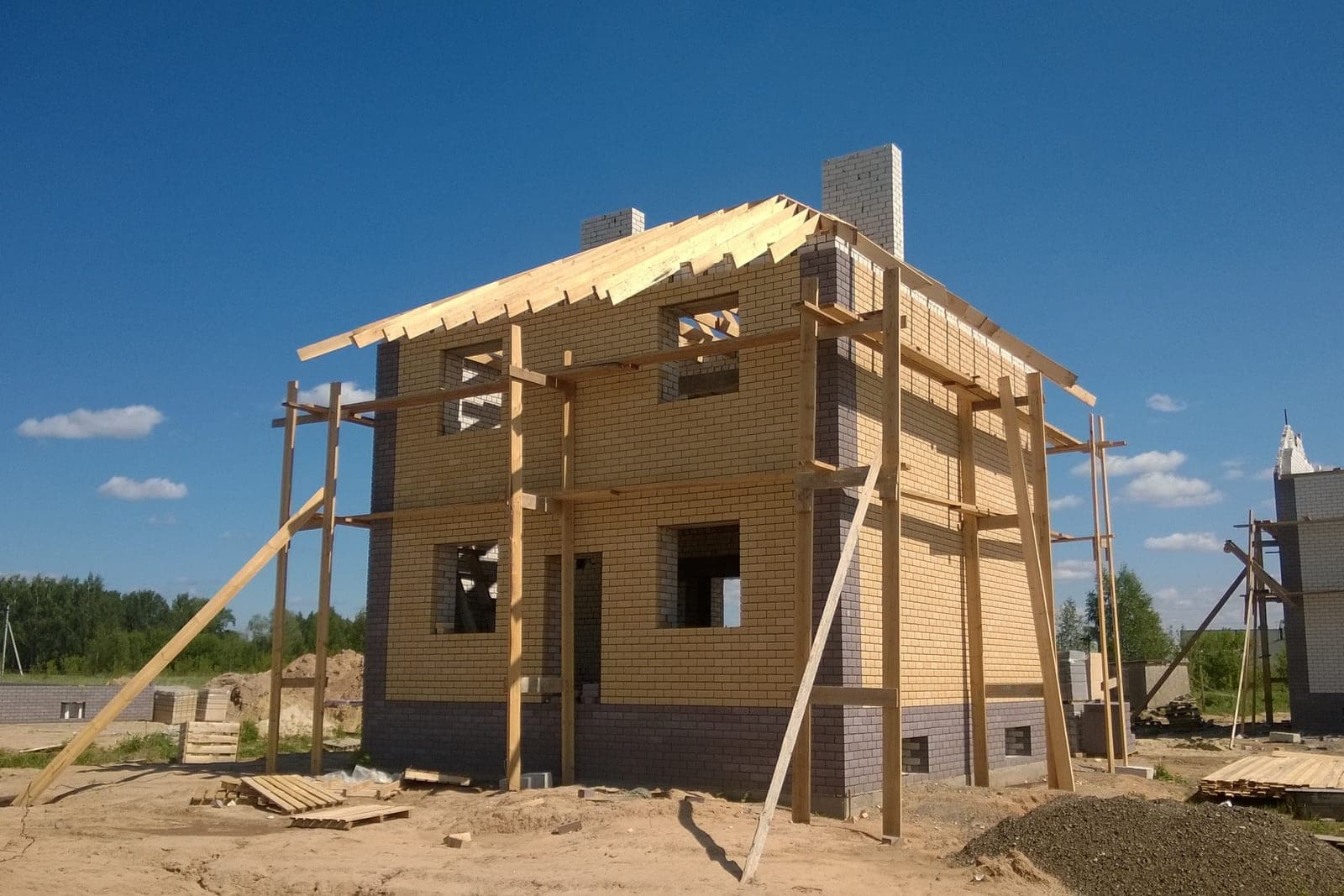 A two-story brick house under construction, with new roof rafters and wooden scaffolding poles against a blue sky