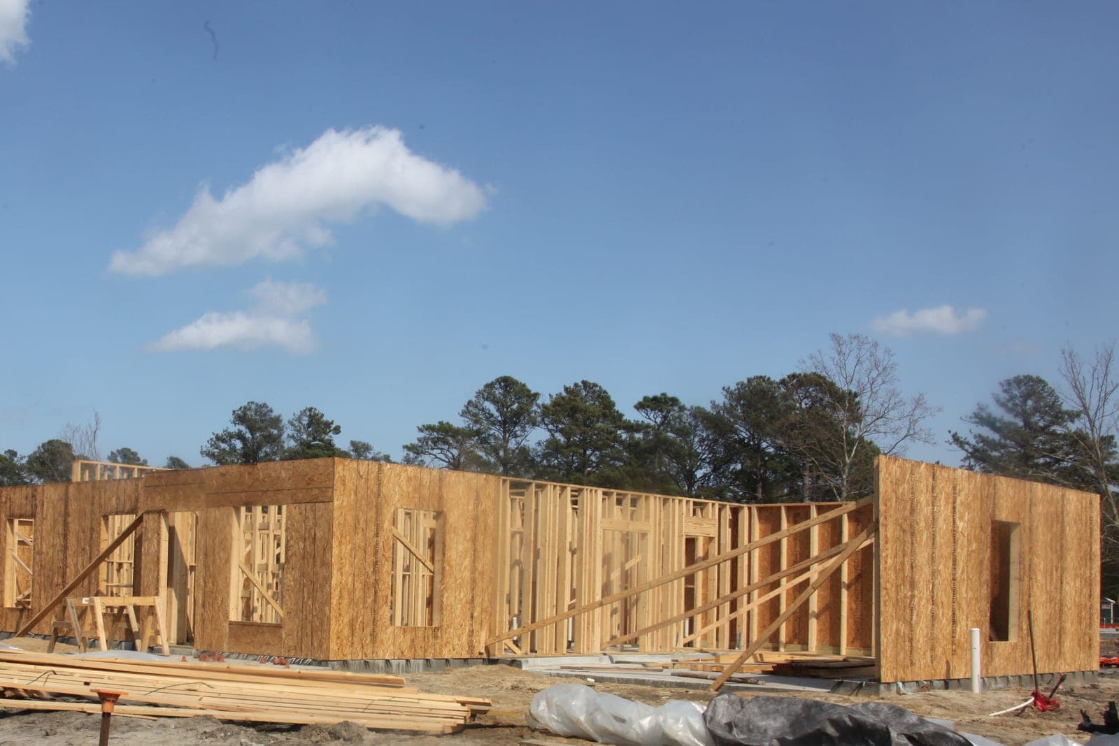 Wood-framed walls of new homes stand on a building site under a blue sky, with stacked lumber in front