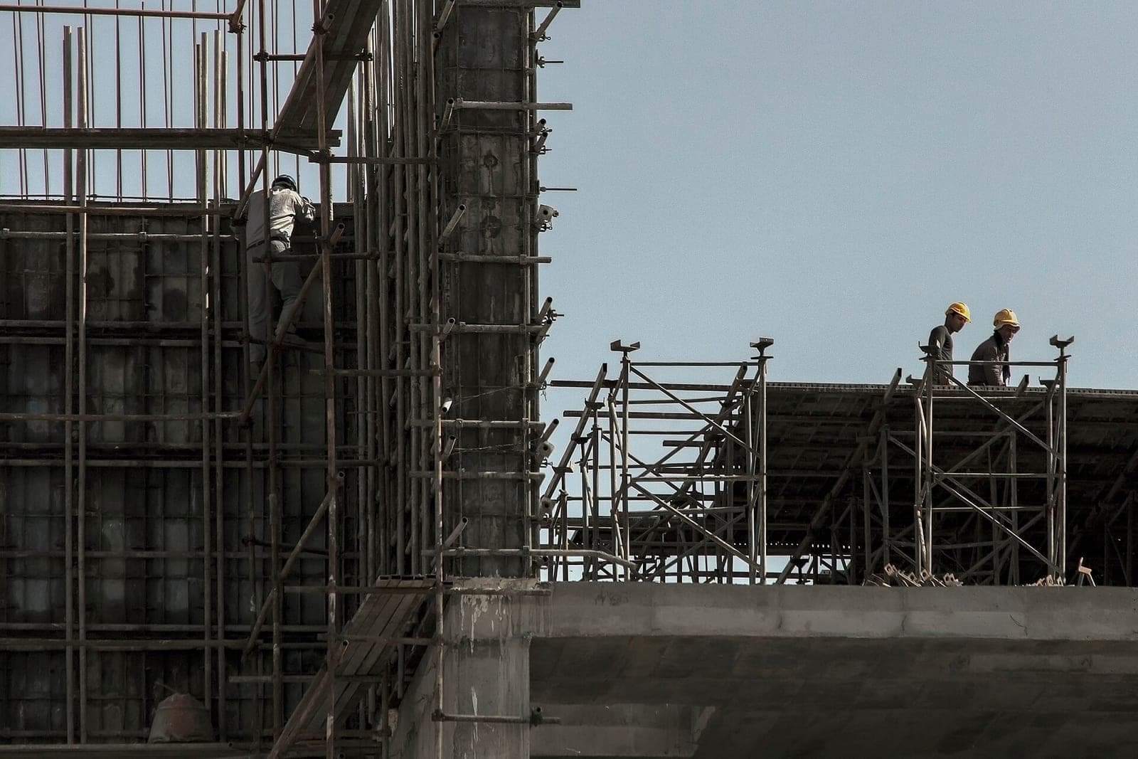 Workers in hard hats walk along scaffolding on top of a concrete building under construction