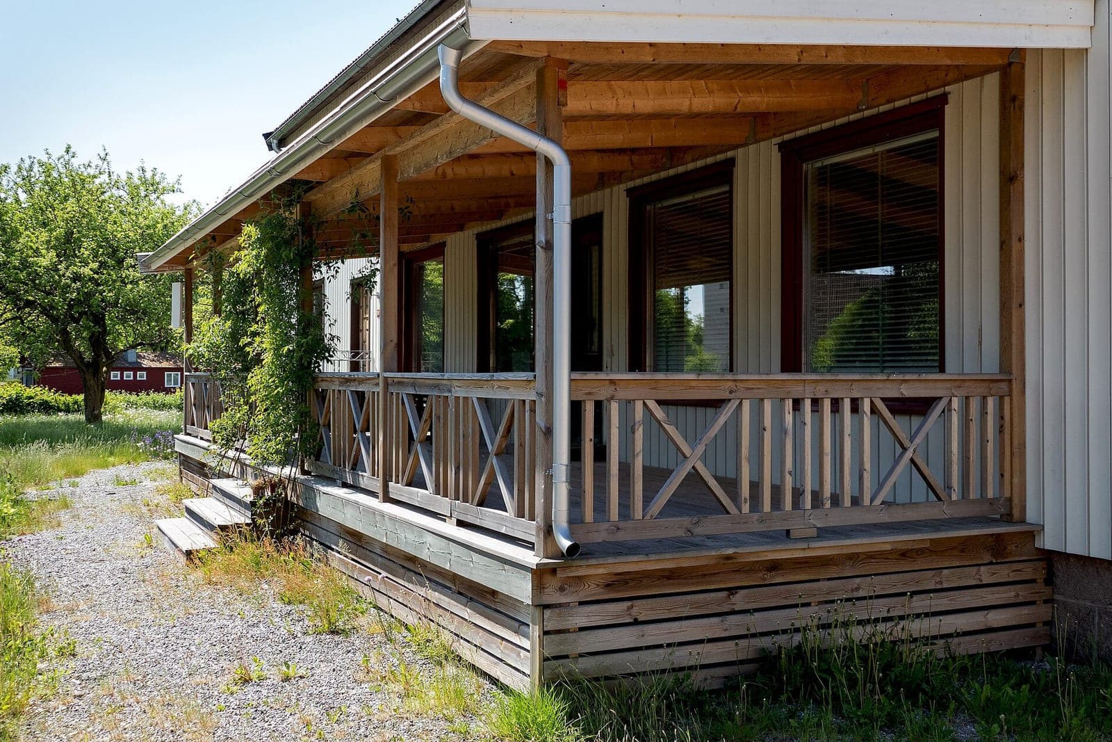 A finished covered wooden porch with crossed railings and a gravel path along a pale wooden house