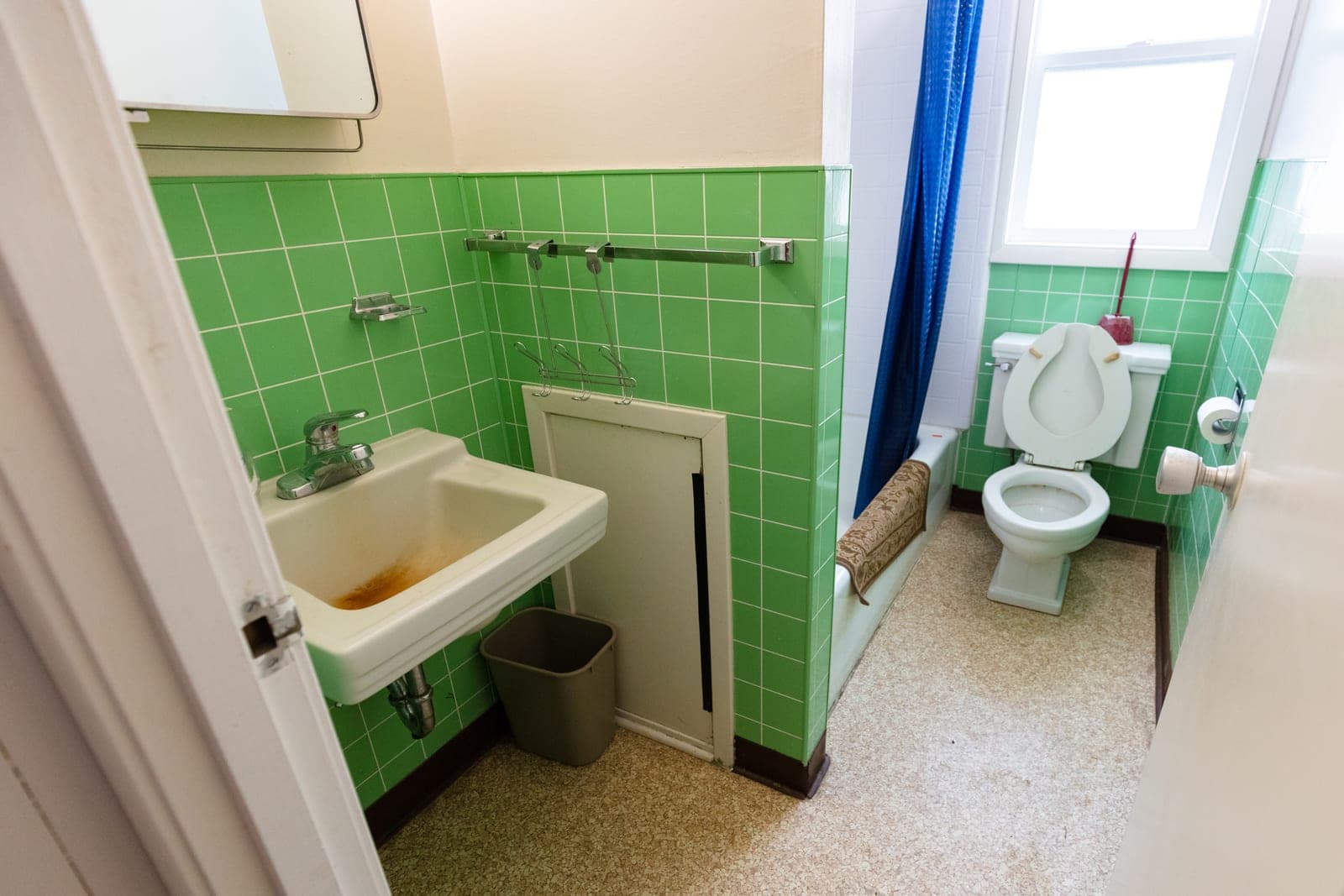 The same small bathroom before its remodel, with green wall tile, a wall-hung sink, a blue shower curtain and an older toilet