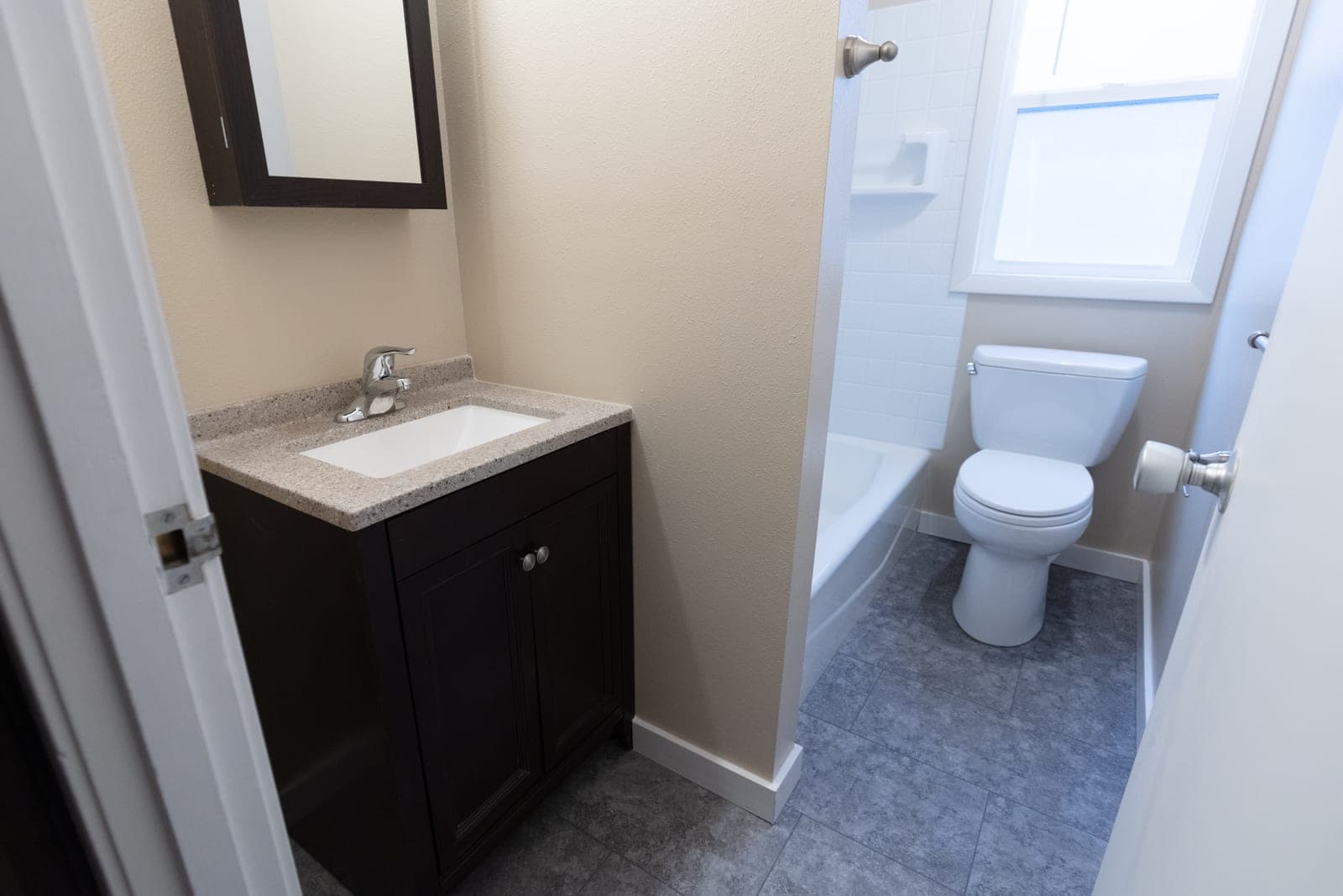 A remodeled bathroom with a dark wood vanity, a new toilet and tub, and a gray tile floor, seen through an open door