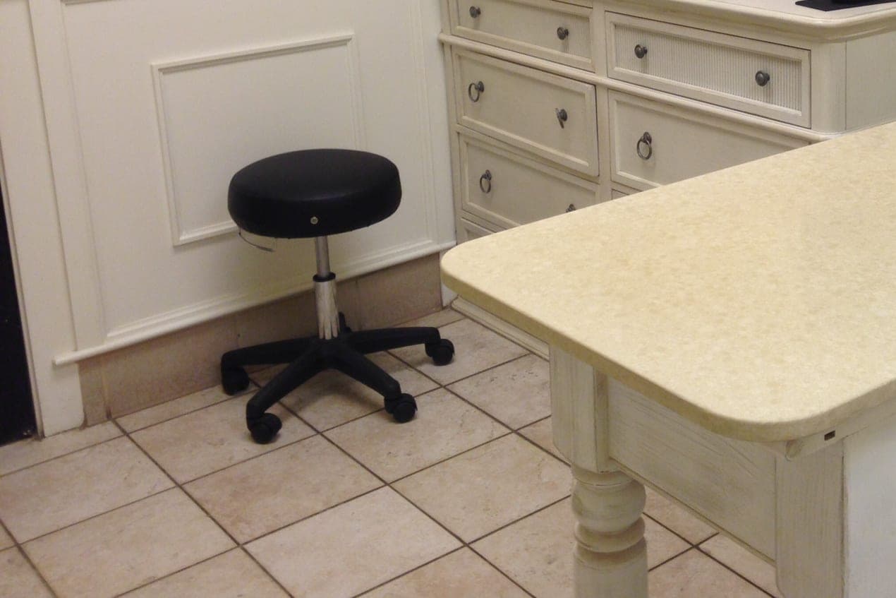 A black rolling stool beside a cream exam table and a chest of drawers on a tiled floor in a veterinary exam room