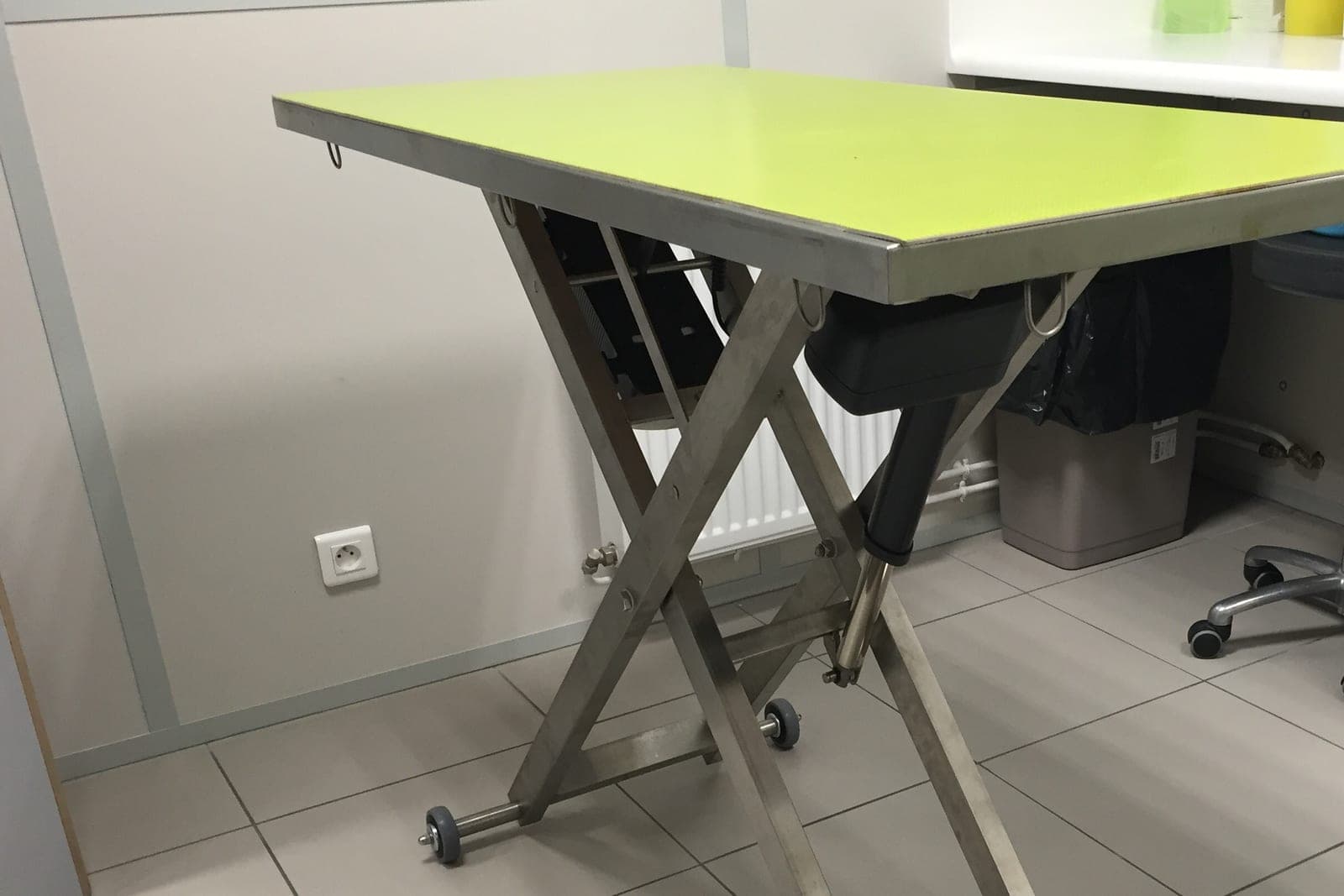 A stainless steel exam table with a bright green top on scissor legs in a tiled veterinary exam room