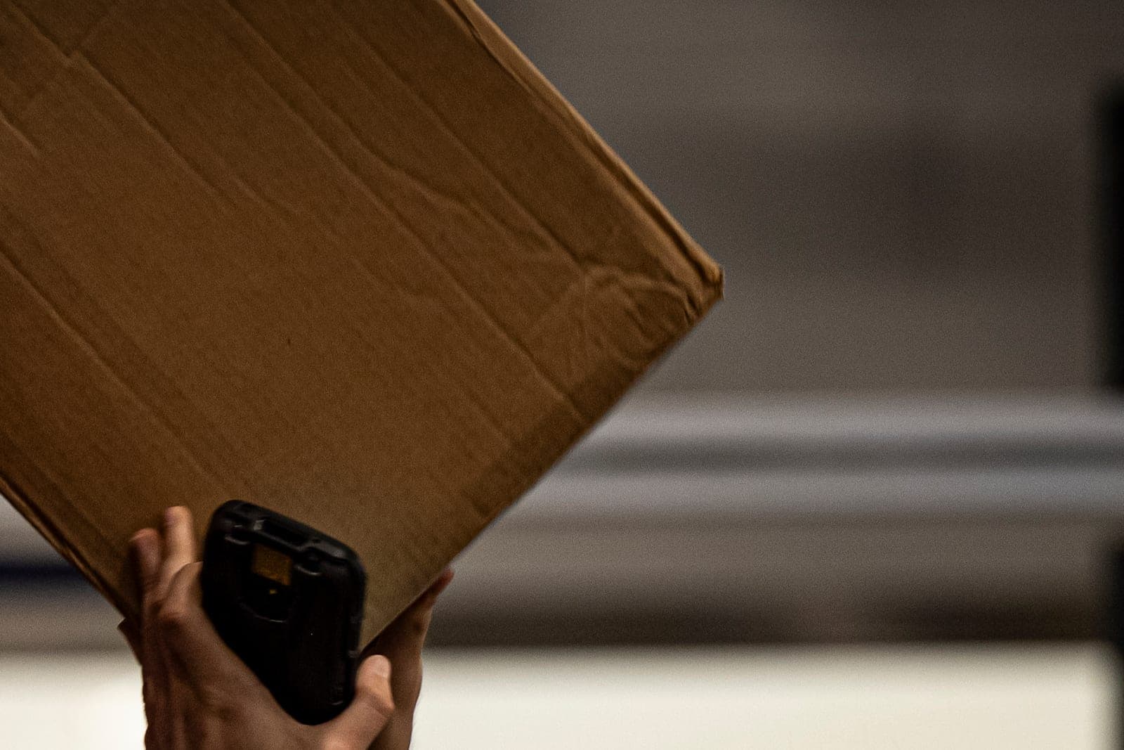 Two raised hands hold a handheld scanner under a tilted cardboard parcel