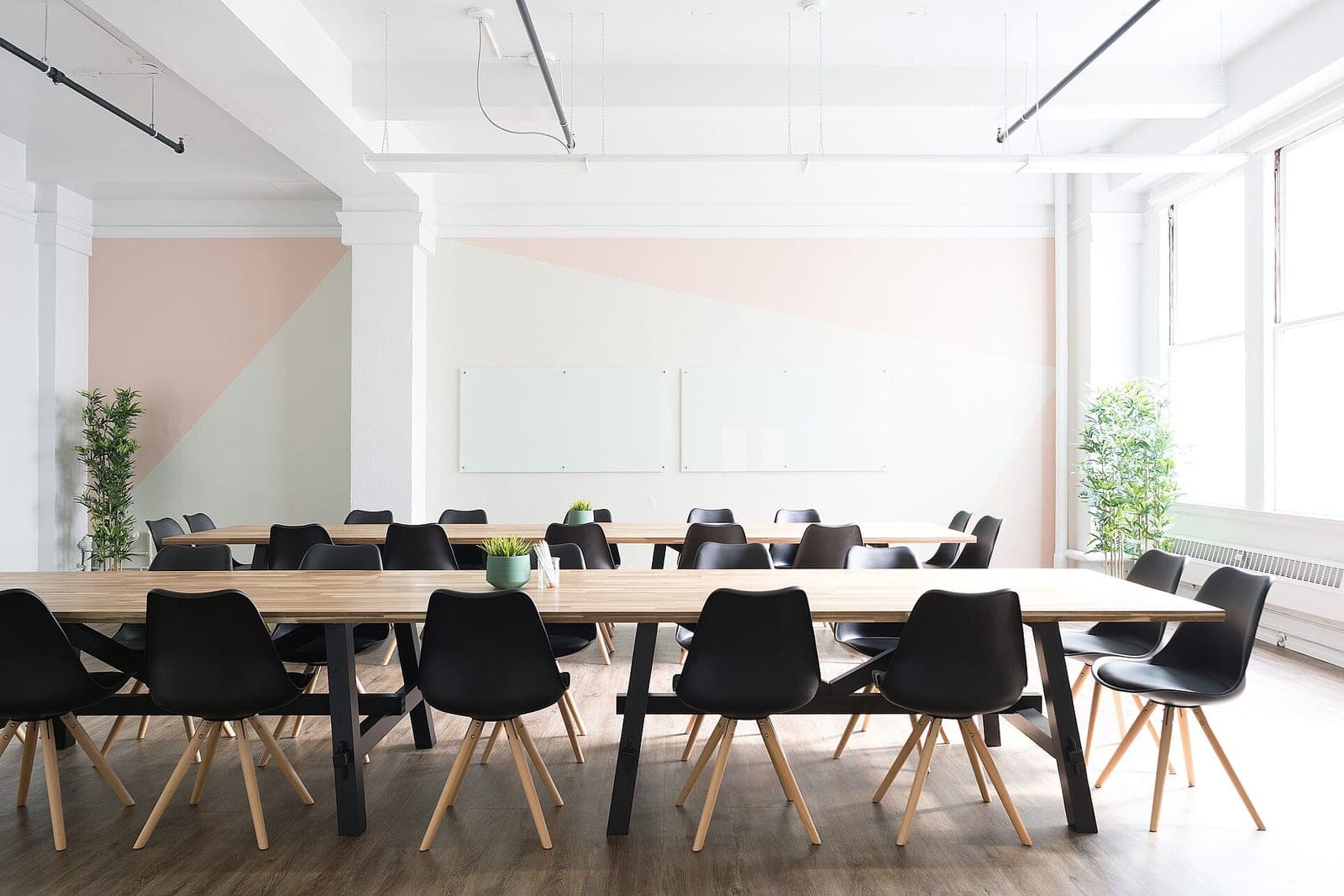 Two long wooden tables with black chairs in a bright meeting room with blank glass boards on a pink and white wall