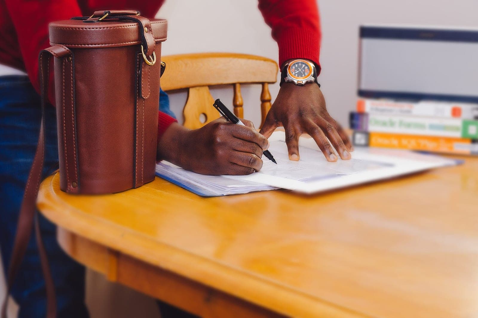A man in a red sweater signs a paper form at a wooden table beside a leather bag