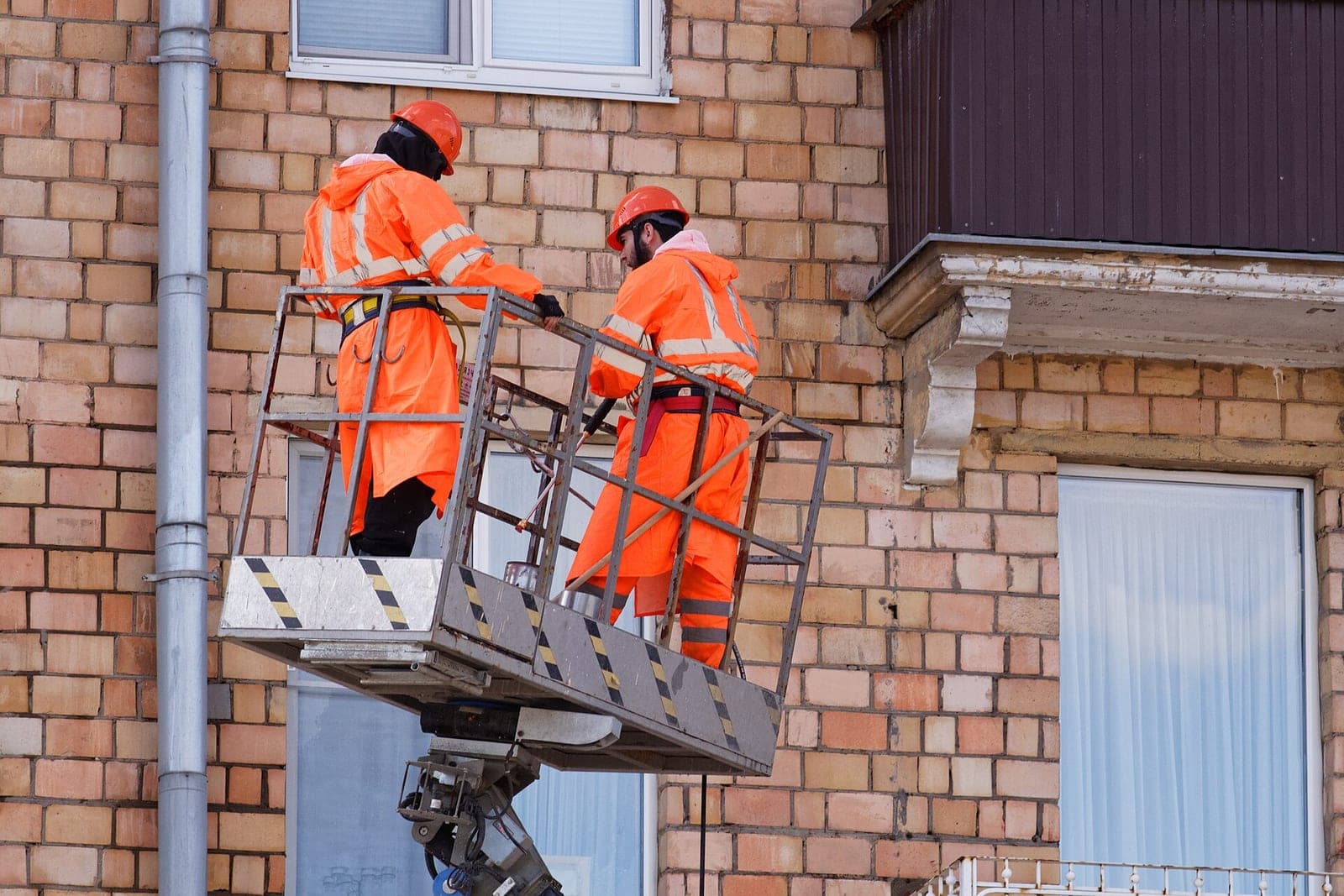 Two workers in orange rain gear wash a brick building wall from a lift basket