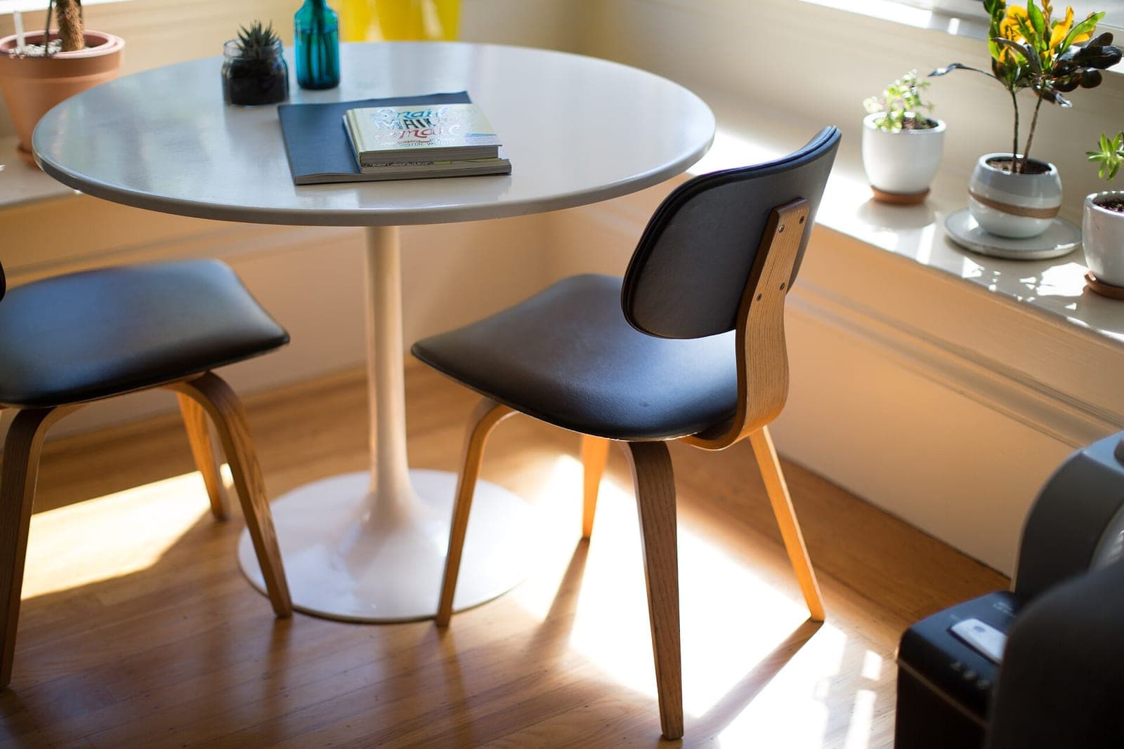 A round white table with two wood and black chairs in a sunny room with potted plants on the windowsill