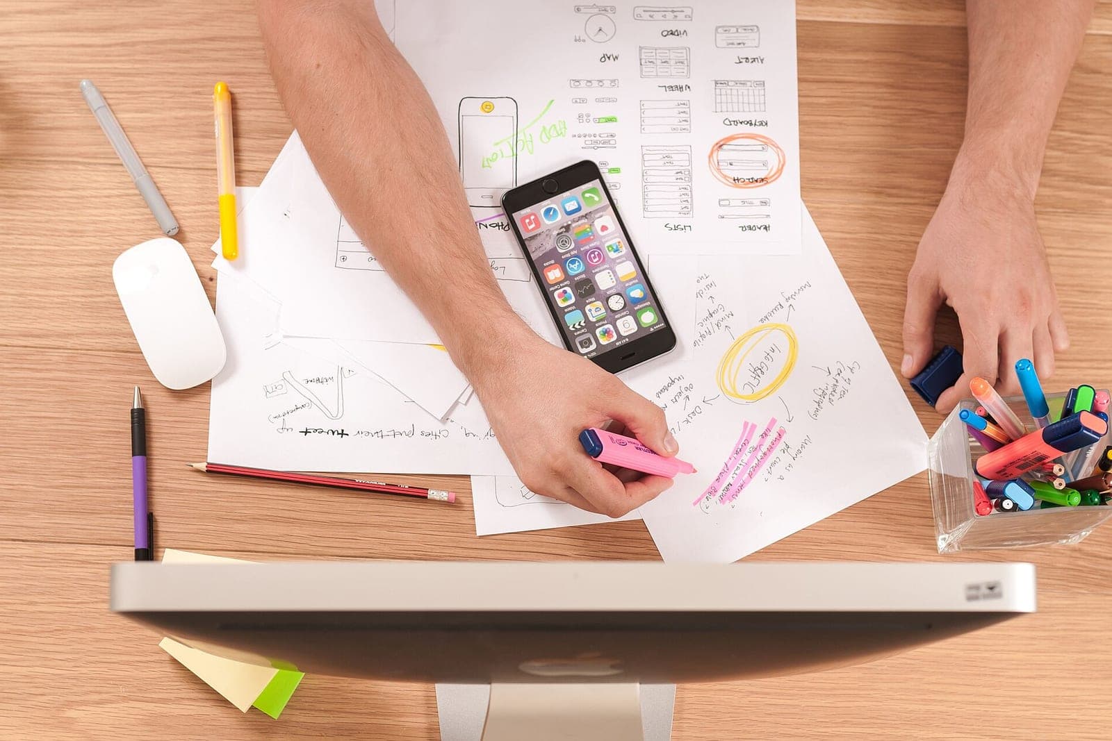 Overhead view of hands marking up paper sketches with a highlighter on a wooden desk, beside a phone and pens