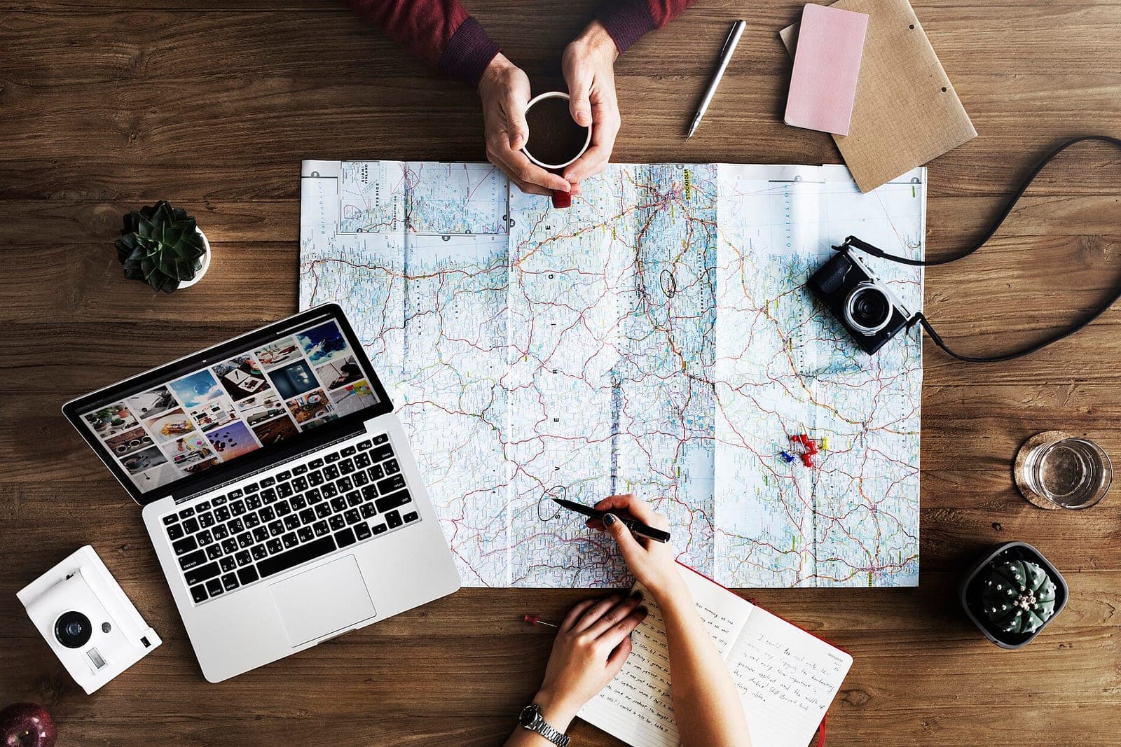 Two people plan a route over a large paper road map on a wooden table, with a laptop, coffee and a camera