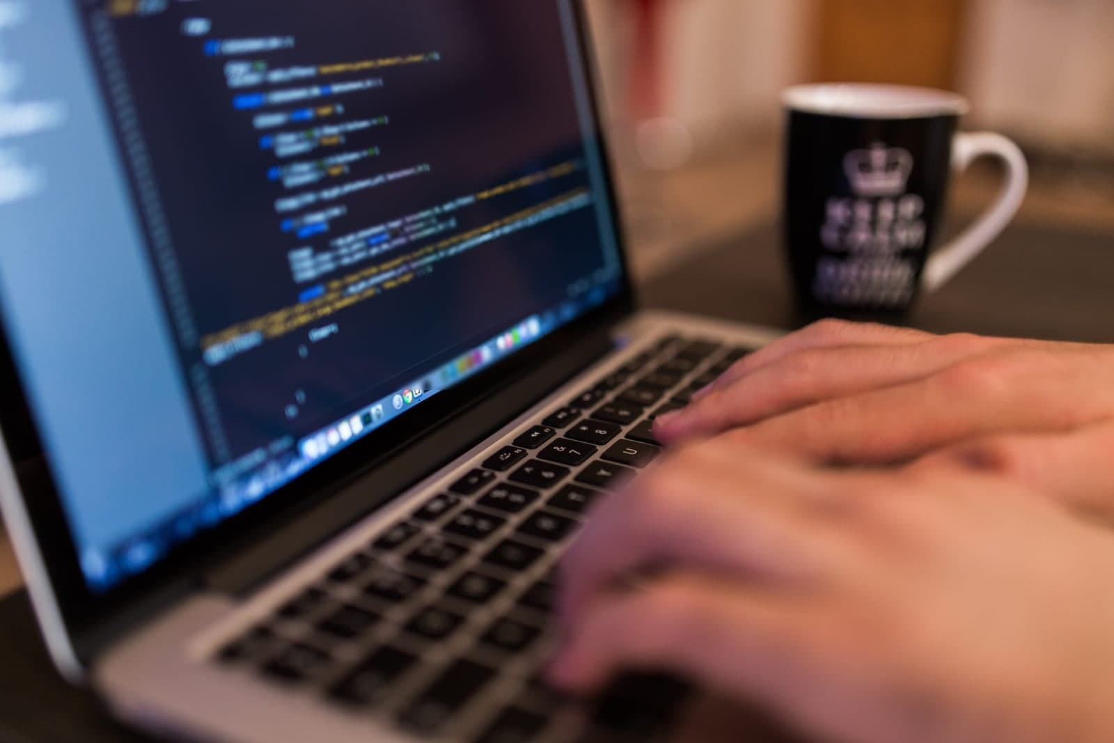 Hands type on a laptop showing lines of code, with a coffee mug on the desk behind