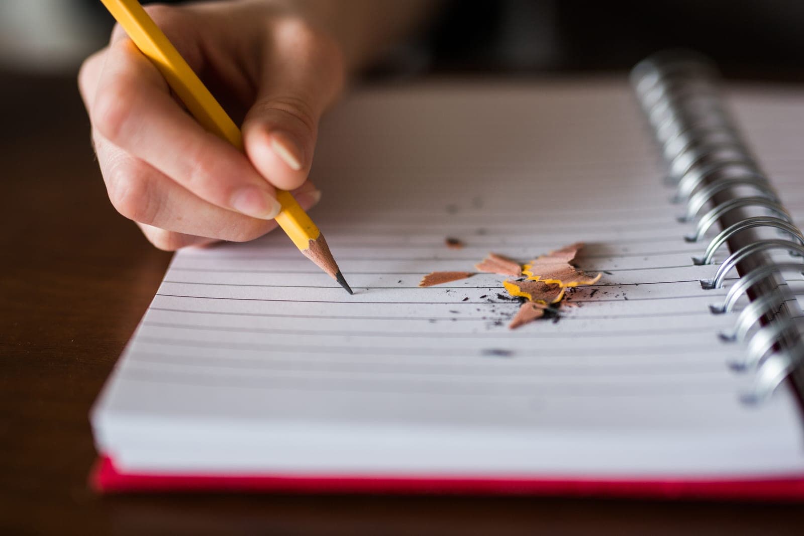 A hand writes with a yellow pencil in a spiral notebook next to pencil shavings.